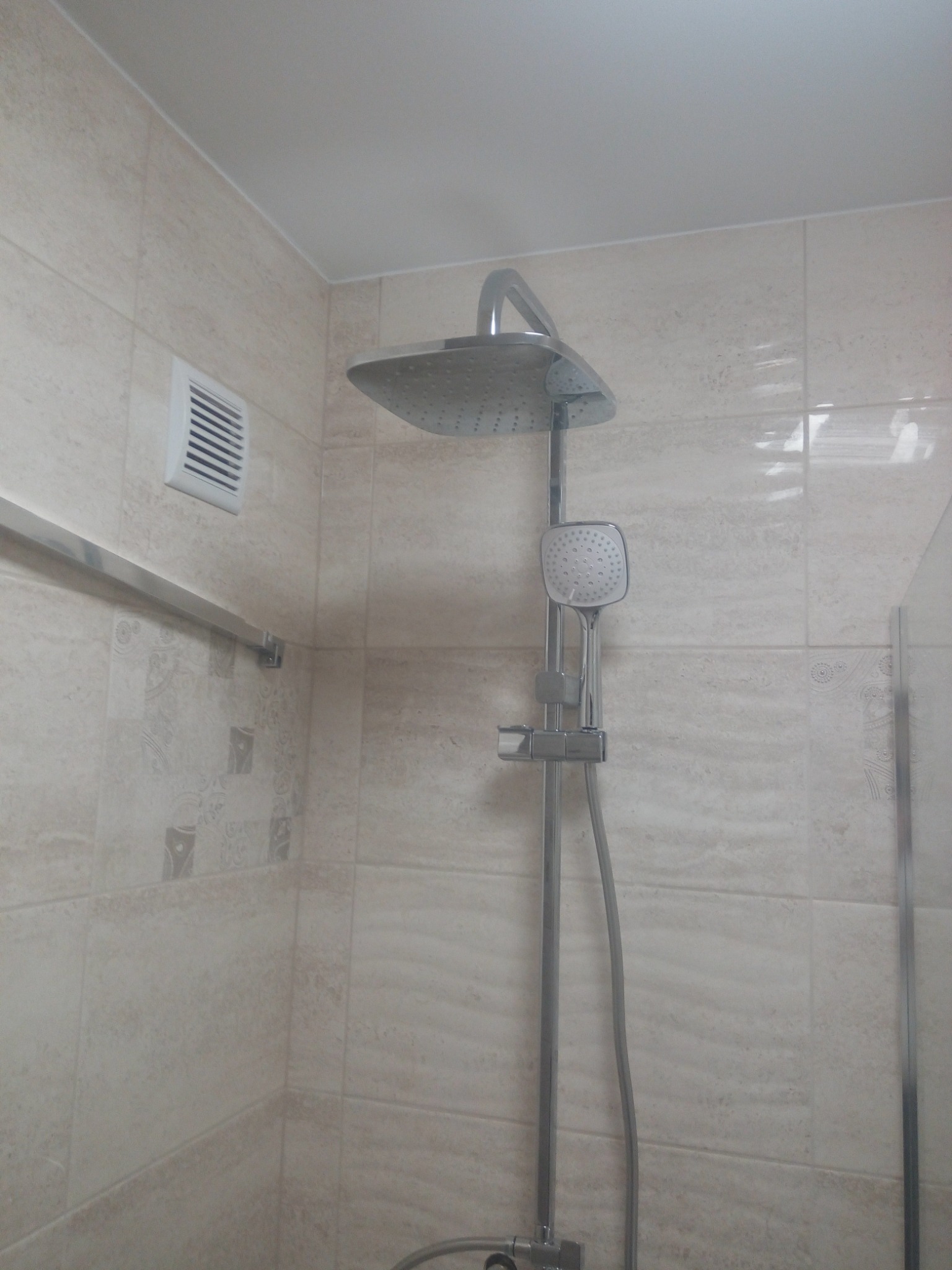Zamontowany zestaw prysznicowy z deszczownicą i słuchawką na tle beżowych płytek w łazience z kratką wentylacyjną.