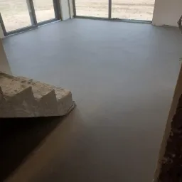 Świeżo wylana, gładka posadzka w surowym wnętrzu z dużymi oknami, widoczne schody w stanie surowym.