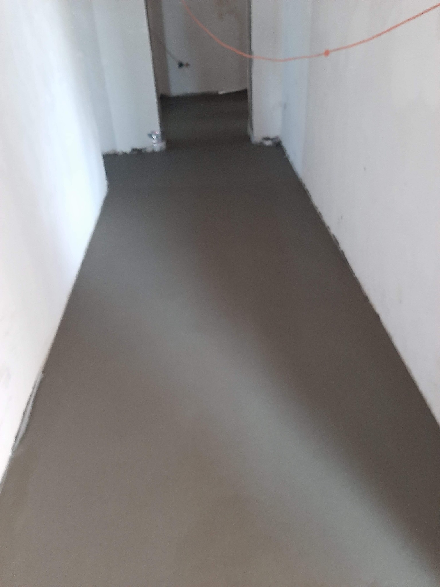 Świeżo wylana, gładka, szara posadzka w pomieszczeniu w trakcie remontu. Na ścianach widoczne ślady po pracach wykończeniowych, a w tle przewody elektryczne.