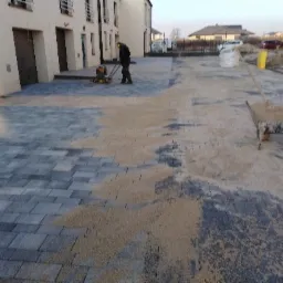 Prace brukarskie w trakcie: zagęszczarka w użyciu na szarej kostce, widoczny wózek wypełniony piaskiem, piła do cięcia kostki oraz budynki w tle.