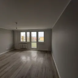 Pusty pok&oacute;j po remoncie z szarymi ścianami, drewnopodobną podłogą, grzejnikiem i oknem z widokiem na zabudowę miejską.