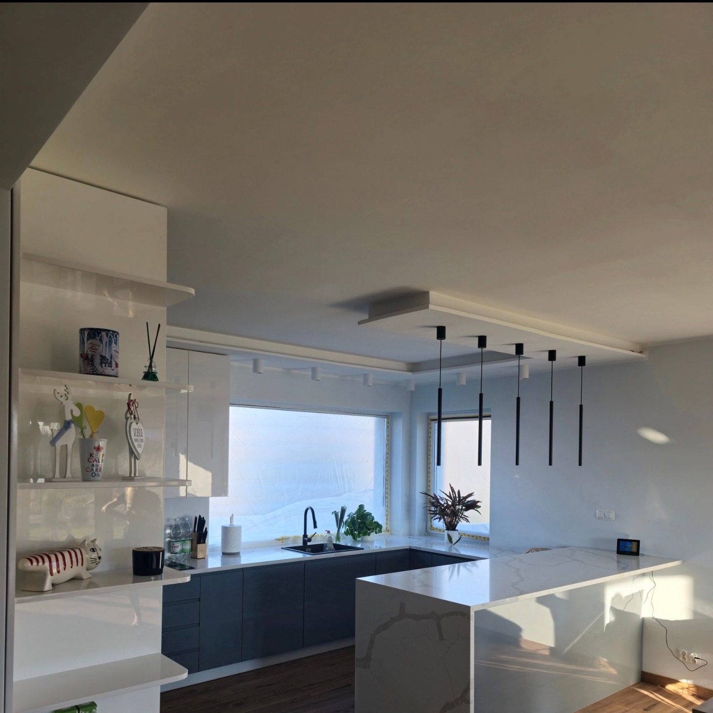 Nowoczesna kuchnia z wyspą i minimalistycznym oświetleniem. Szare szafki, białe blaty, dekoracje na półkach. Duże okno z widokiem na zewnątrz.