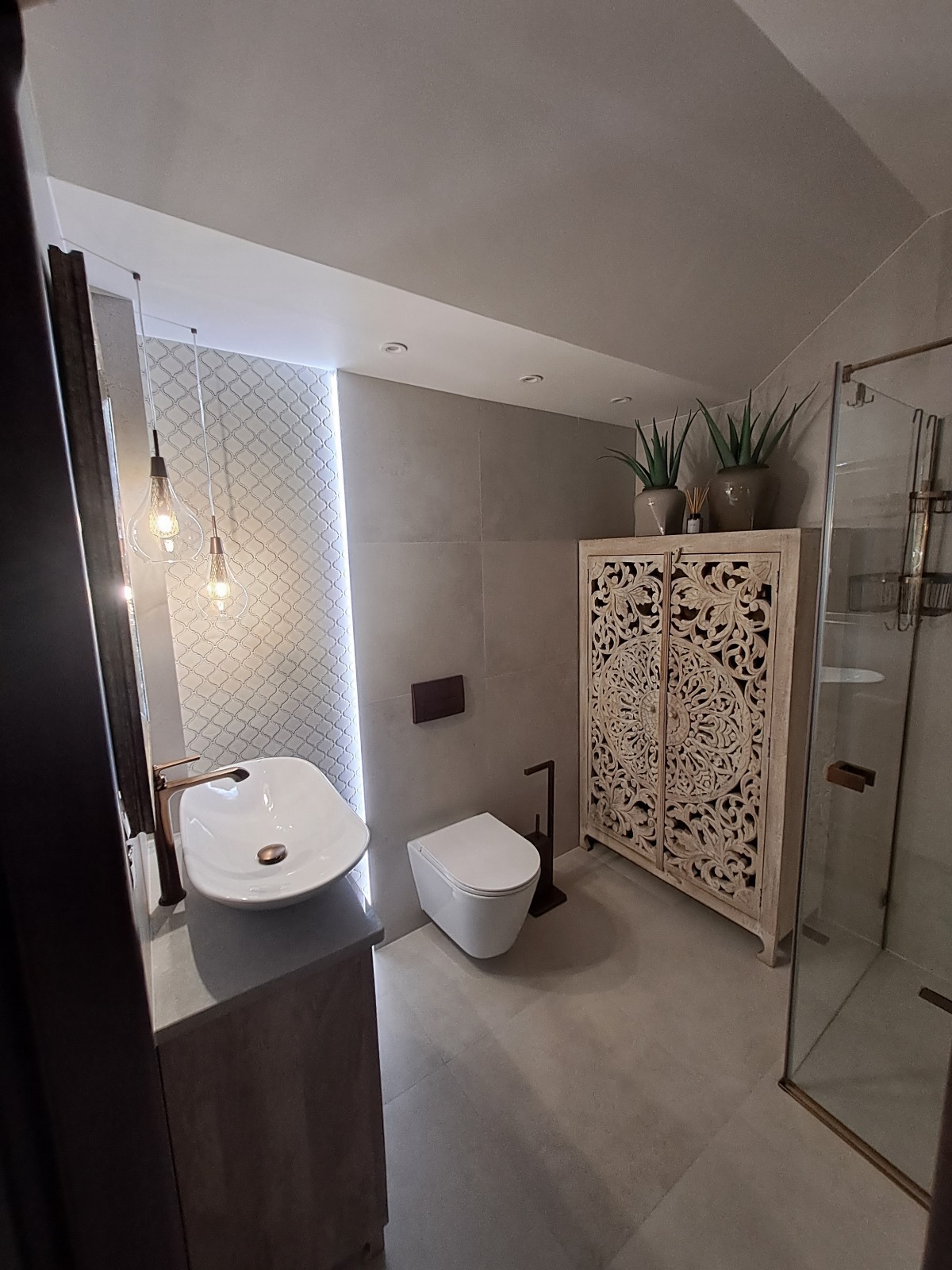 Jasna łazienka z umywalką na blacie, toaletą i zdobioną szafką. Oświetlenie punktowe i dekoracyjne rośliny doniczkowe dodają wnętrzu charakteru.