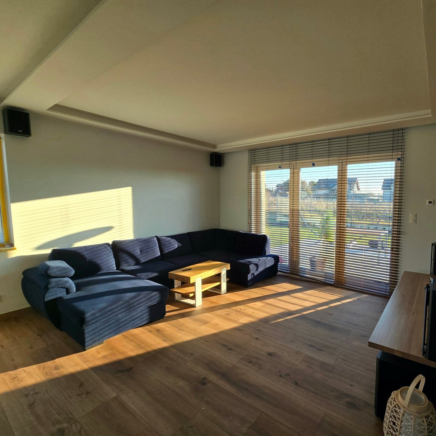 Salon z drewnianą podłogą, narożnikiem w odcieniach granatu i stolikiem kawowym. Promienie słońca wpadają przez okno z żaluzjami, oświetlając wnętrze.