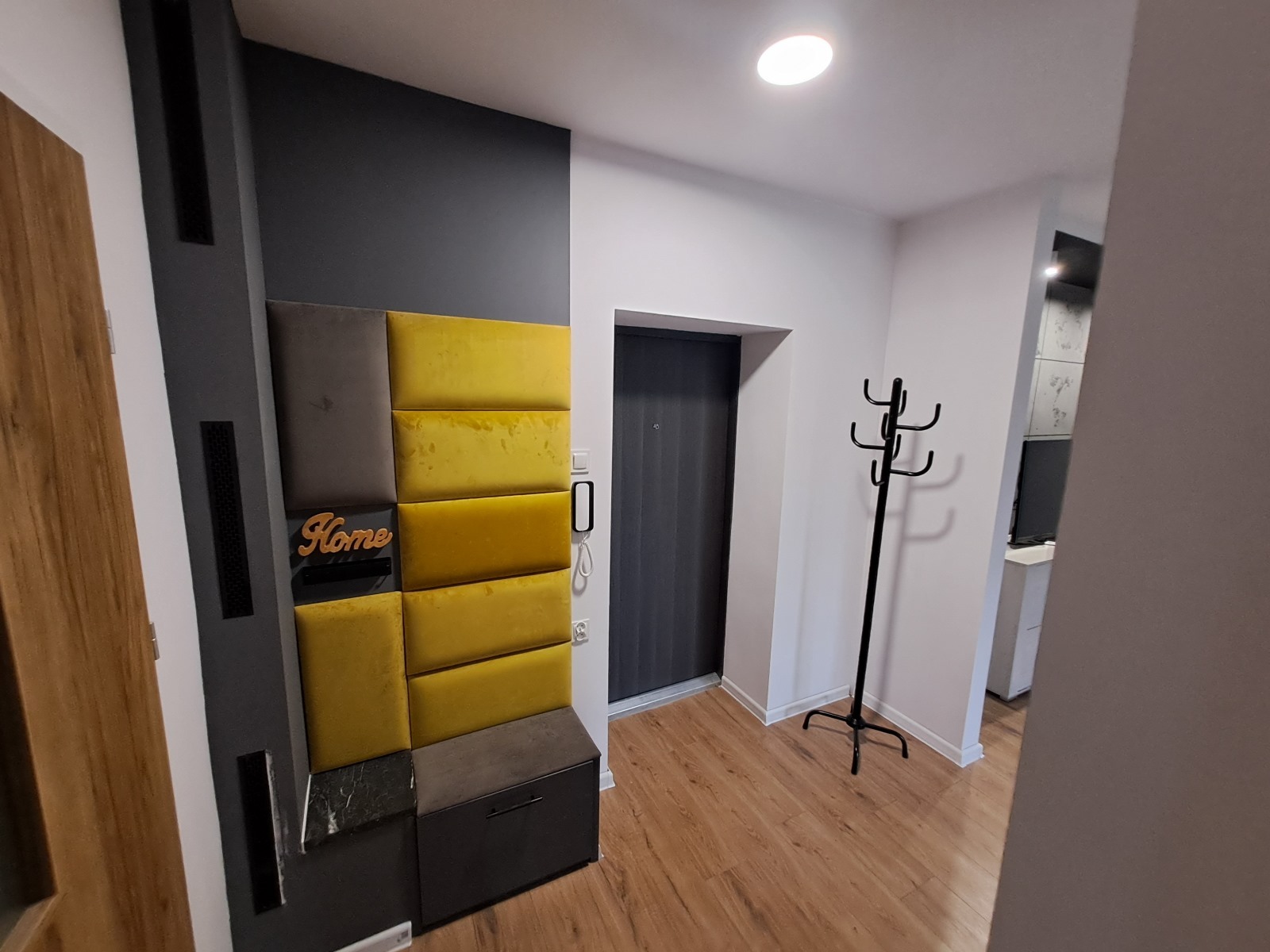 Nowoczesny przedpokój z panelami ściennymi w odcieniach szarości i żółci, drewnianą podłogą i czarnym wieszakiem. Widoczna zabudowa z siedziskiem i szafką.