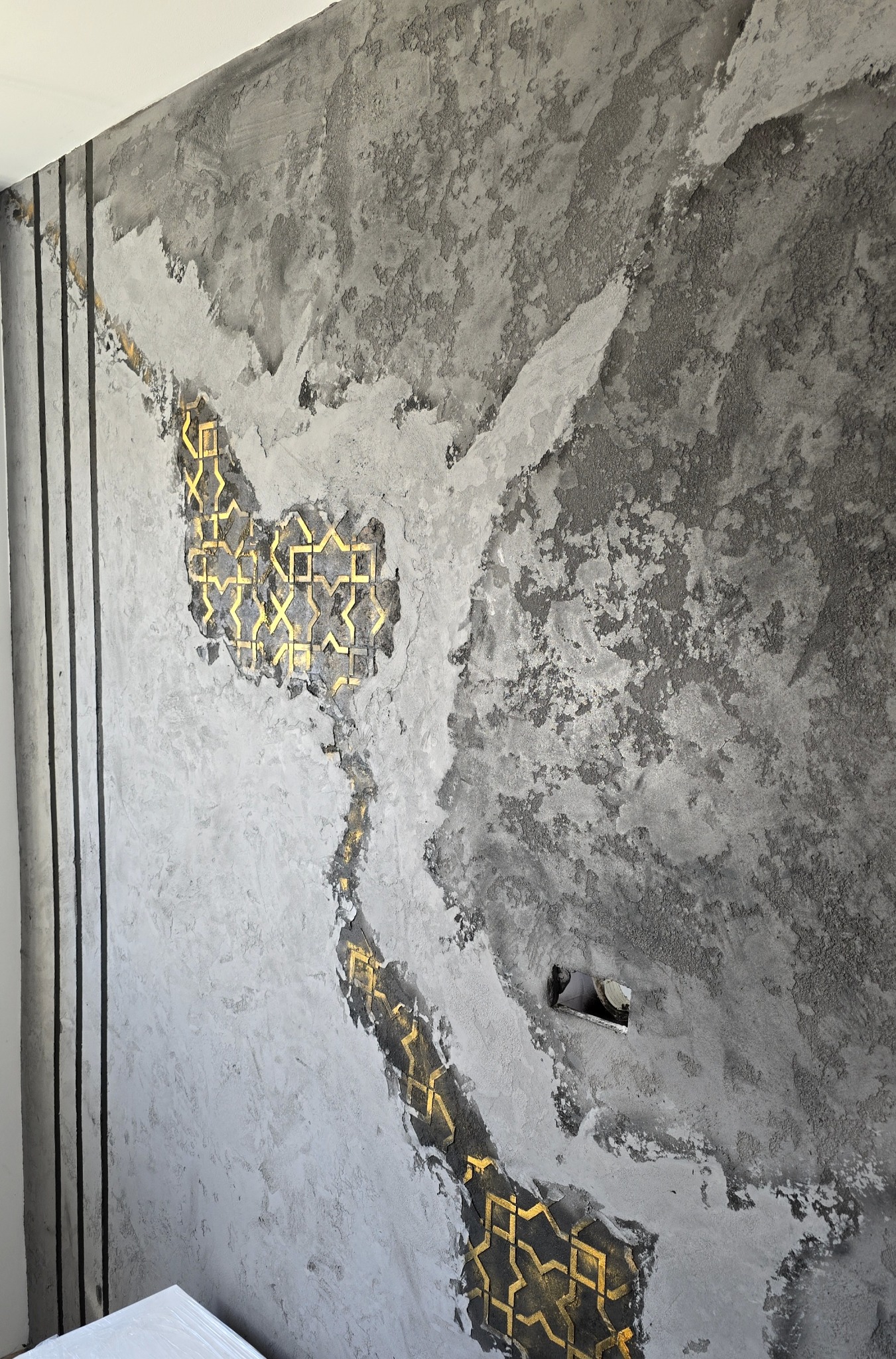 Dekoracyjna ściana z efektem surowego betonu, wzbogacona o złote, geometryczne ornamenty i pionowe, czarne listwy. Widoczna puszka elektryczna.