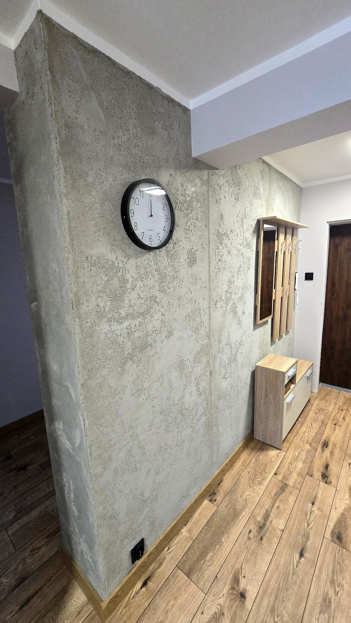 Wykończony przedpokój z dekoracyjną ścianą w odcieniach szarości, drewnianą podłogą i minimalistycznymi meblami. Na ścianie wisi zegar i wieszak z lustrem.