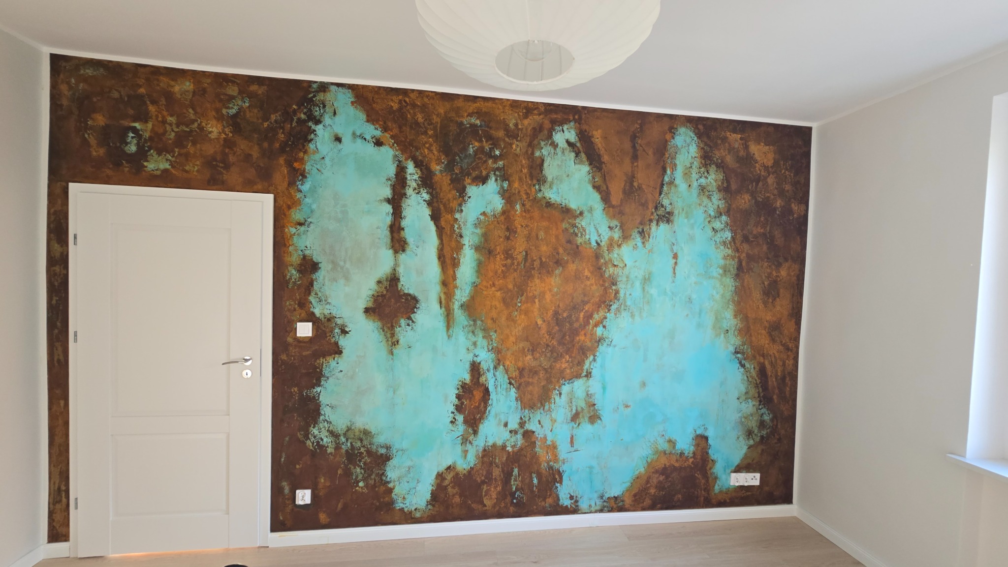 Ściana z dekoracyjną, abstrakcyjną fakturą w odcieniach brązu i turkusu, oświetlona lampą sufitową. Widoczna biała stolarka drzwiowa i jasna podłoga.
