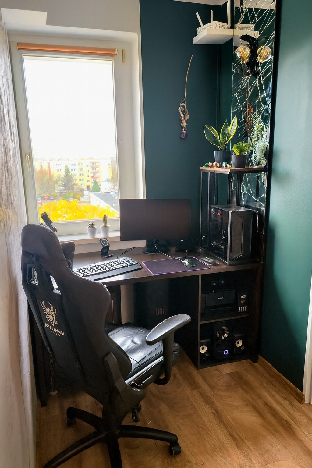 Pokój gracza z biurkiem, komputerem, fotelem gamingowym i dekoracjami. Ściana w kolorze butelkowej zieleni, drewniana podłoga i widok z okna na miasto.