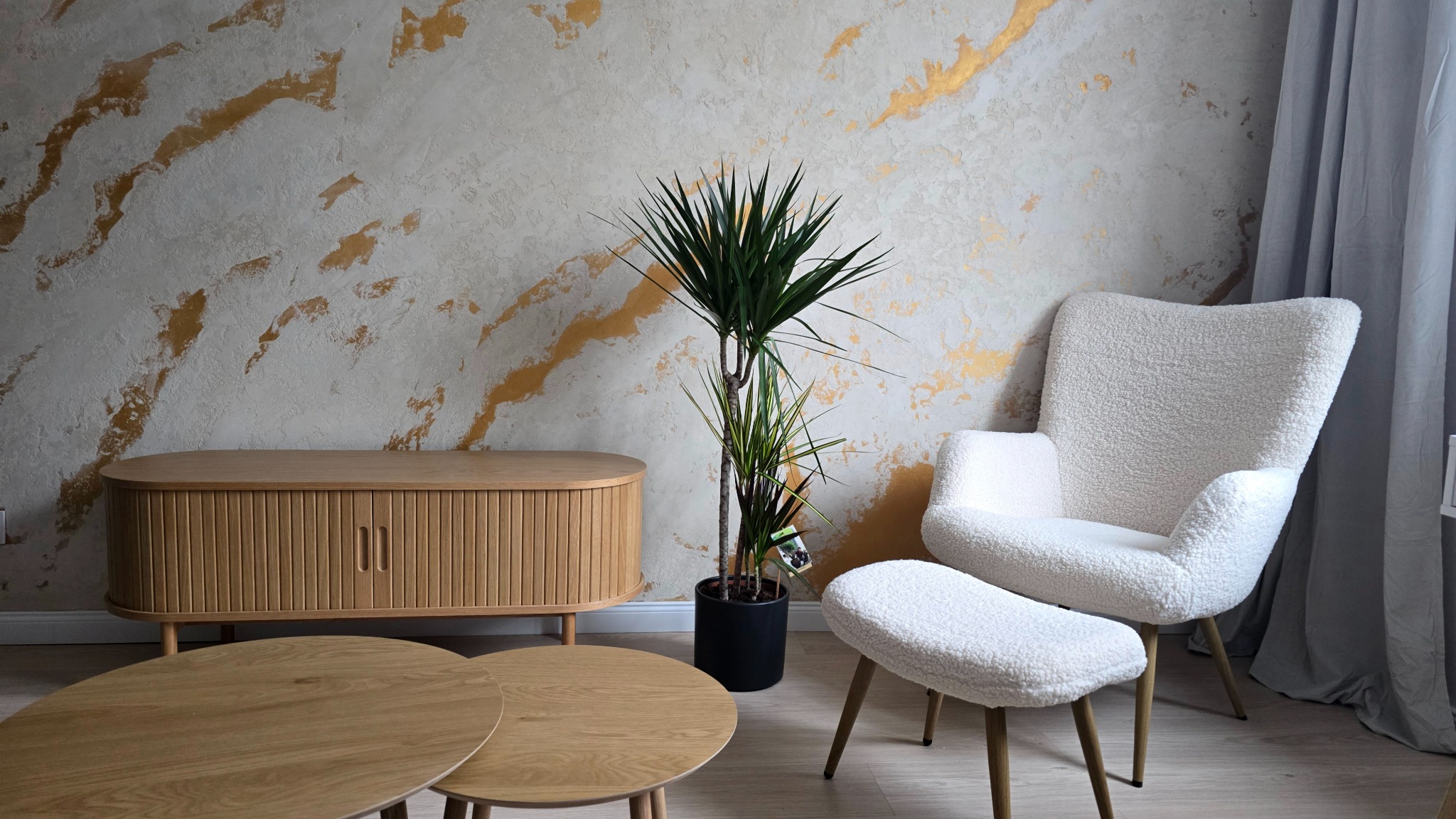 Jasne wnętrze z dekoracyjną ścianą w odcieniach bieli i złota. Drewniana komoda, stoliki kawowe i fotel z podnóżkiem tworzą przytulną aranżację.