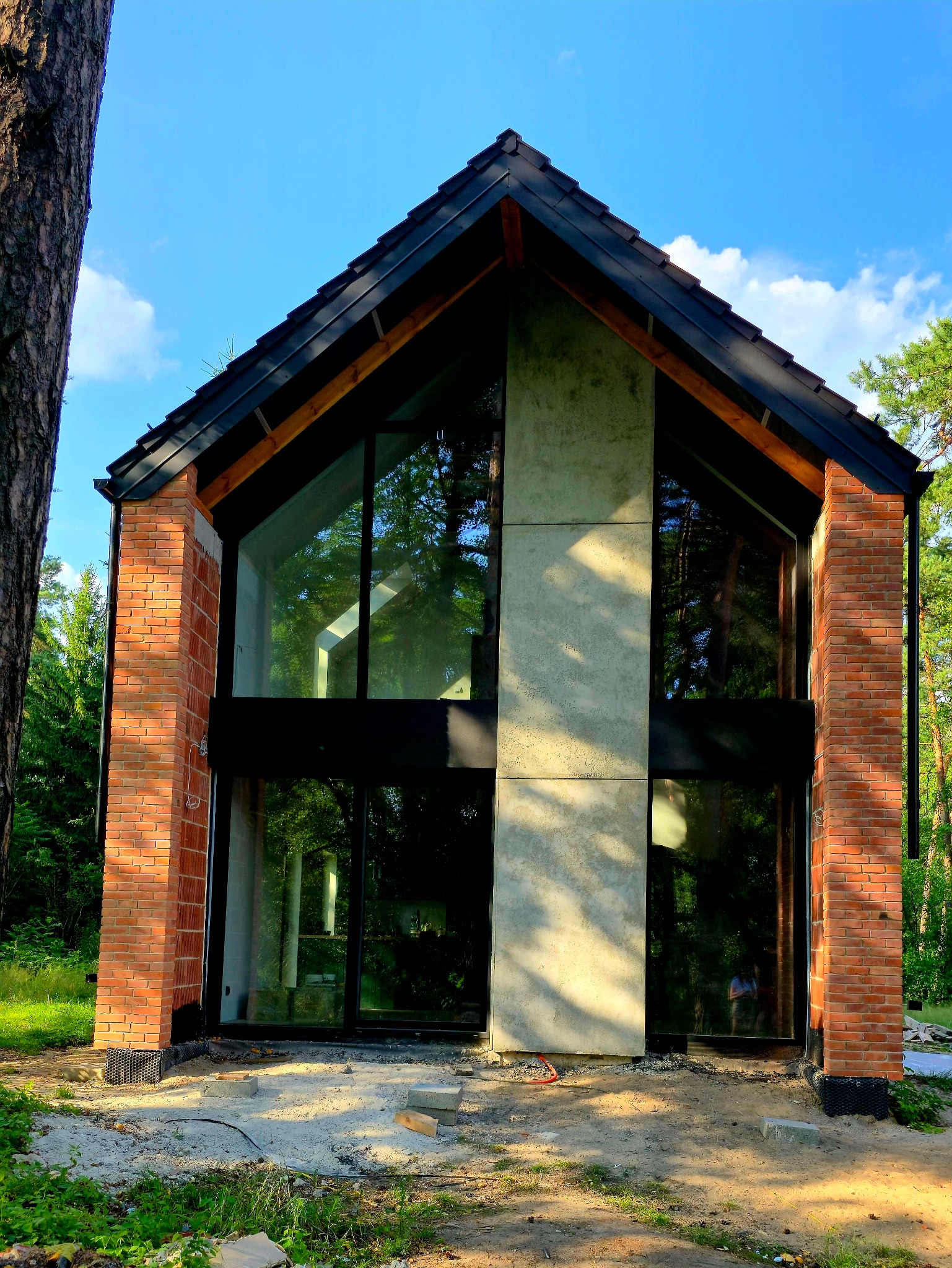 Nowoczesny dom z ceglaną elewacją i dużymi przeszkleniami, otoczony zielenią lasu. Widoczne detale wykończenia i surowy stan otoczenia.