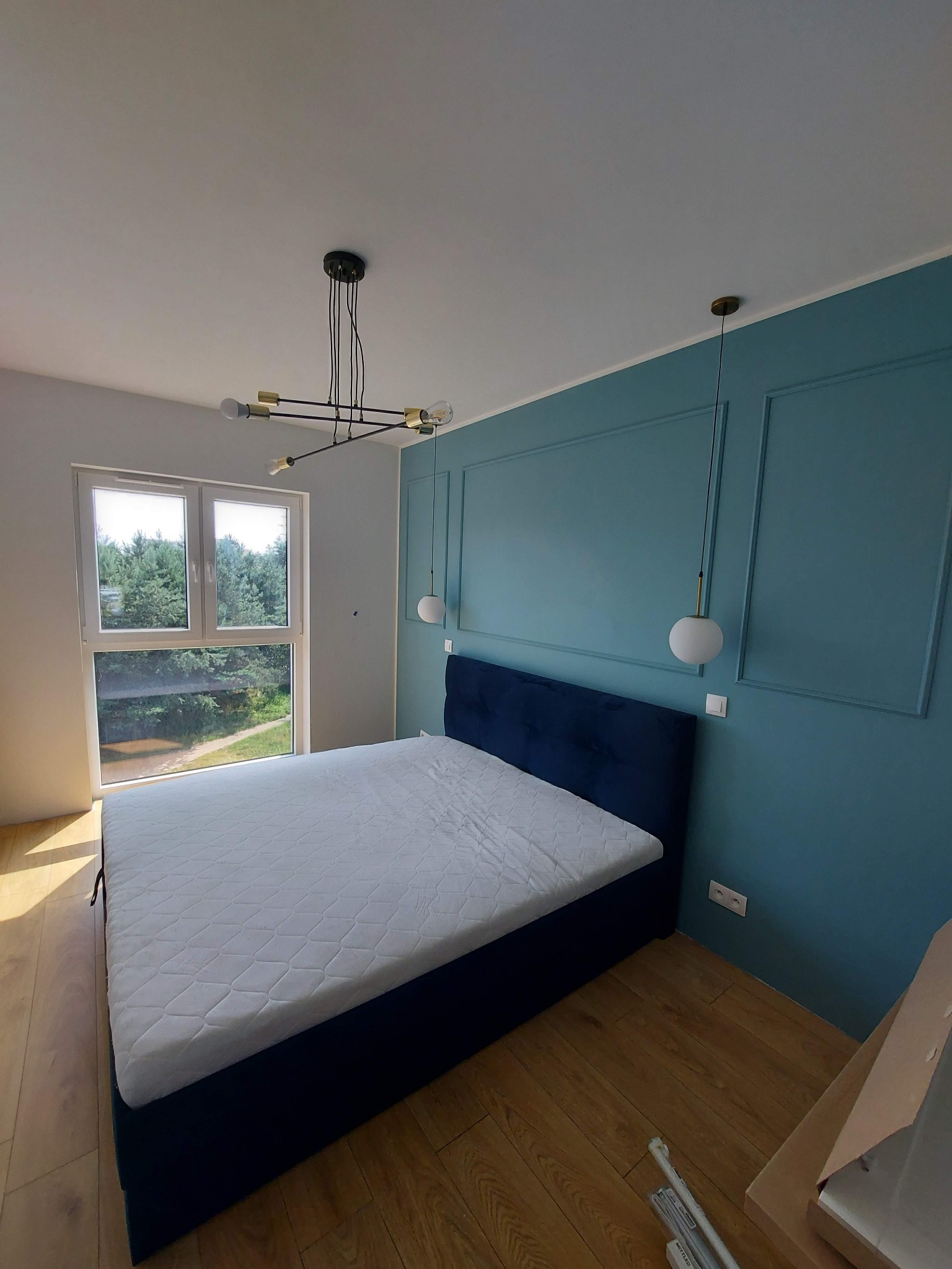 Sypialnia z niebieską ścianą z dekoracyjnymi listwami, łóżkiem z ciemnoniebieskim zagłówkiem i białym materacem, nowoczesnym oświetleniem sufitowym oraz drewnianą podłogą w trakcie wykończenia.