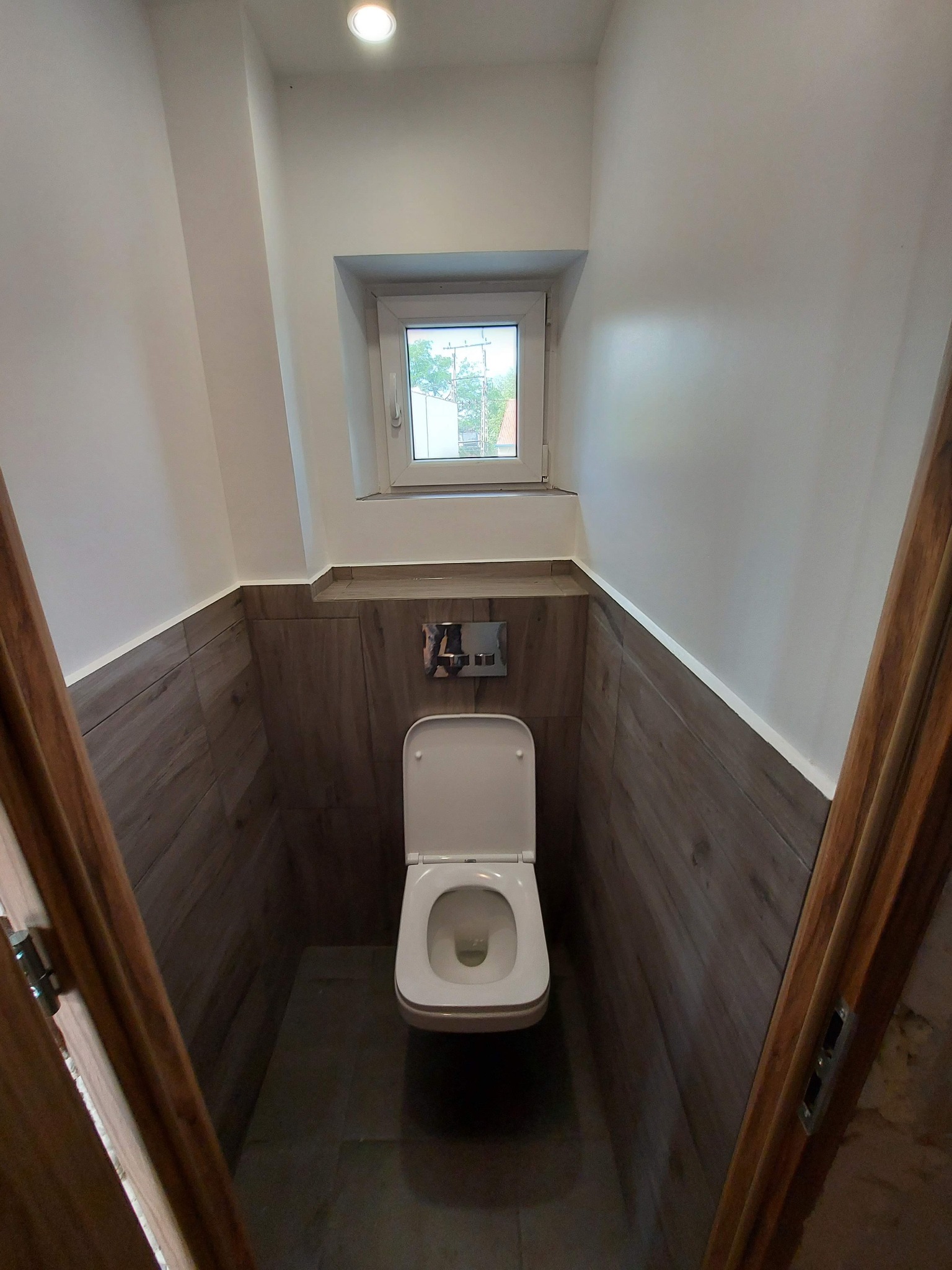 Wąska łazienka z toaletą, wykończona płytkami imitującymi drewno, małe okno z widokiem na zieleń, oświetlenie punktowe w suficie i drewniane futryny drzwiowe.