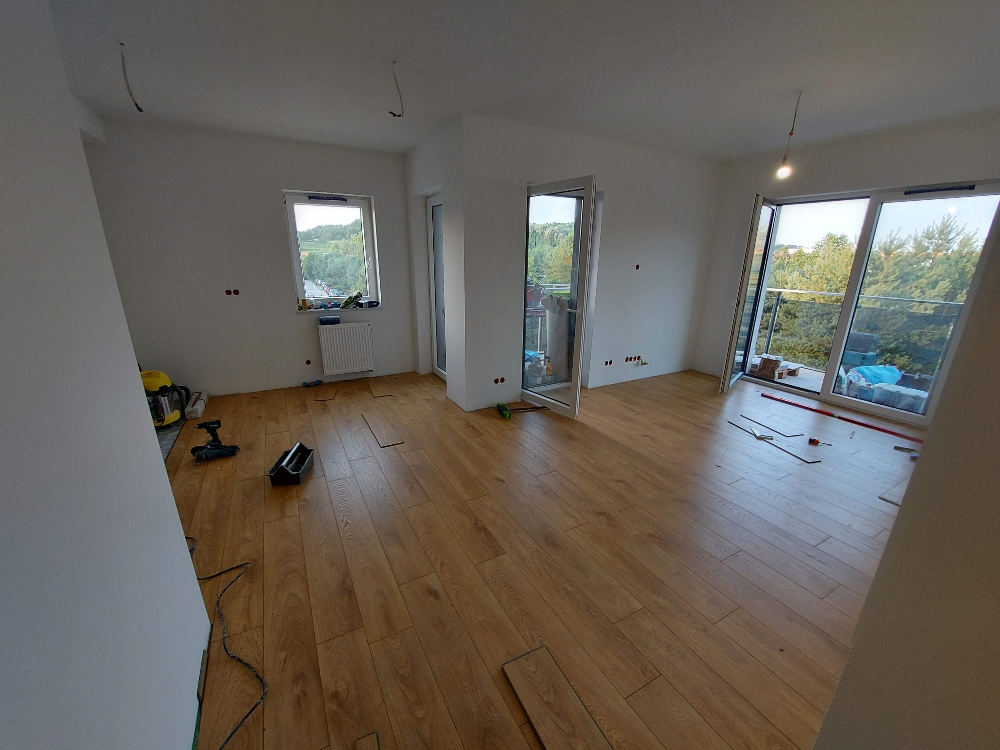 Układanie paneli podłogowych w jasnym, przestronnym pokoju z balkonem, widoczne narzędzia pracy i częściowo ułożona podłoga z paneli drewnopodobnych.