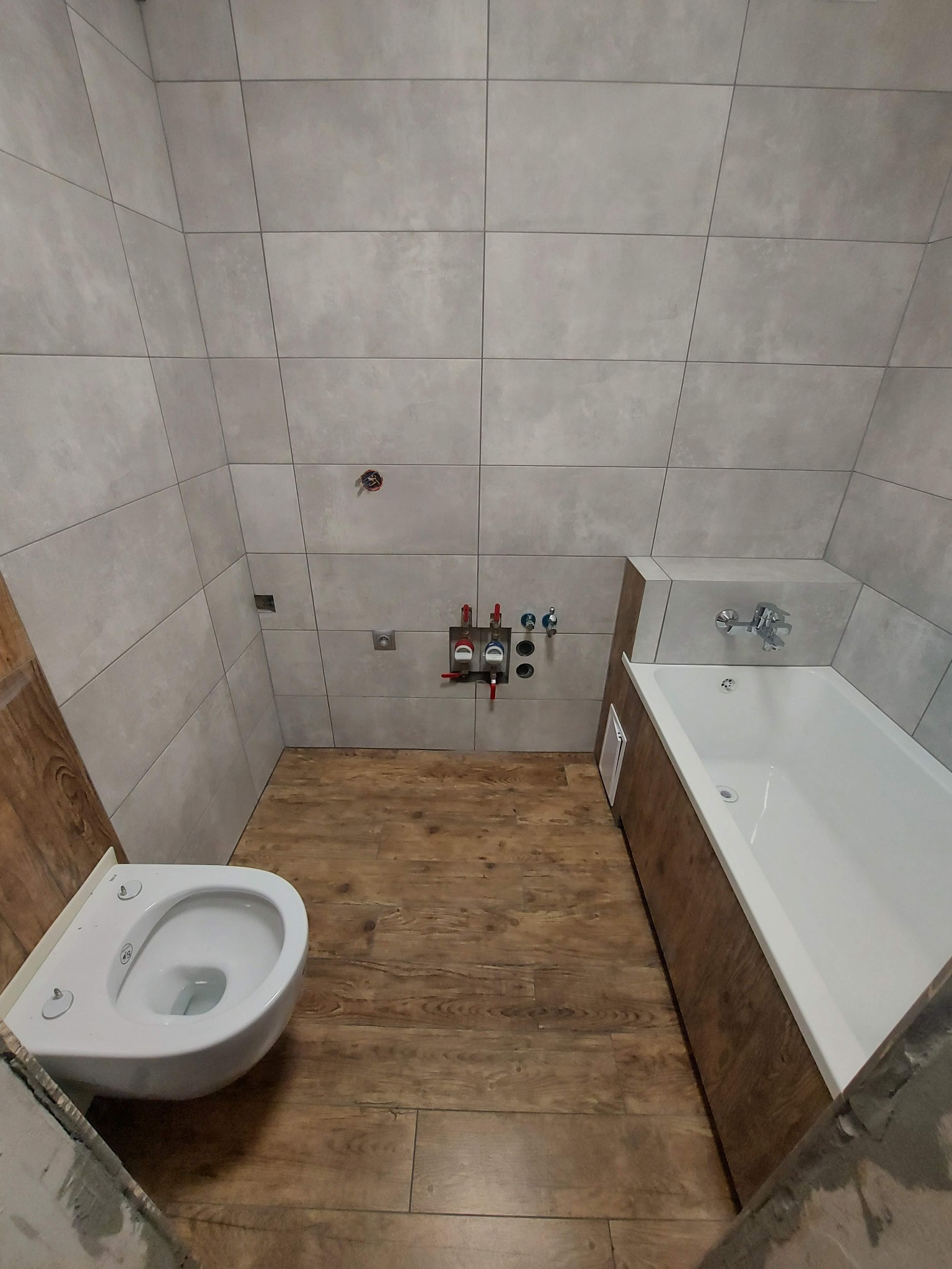 Wnętrze łazienki w trakcie prac wykończeniowych: szare płytki ścienne, drewnopodobna podłoga, zamontowana toaleta, zabudowana wanna z baterią, widoczne instalacje wodne.