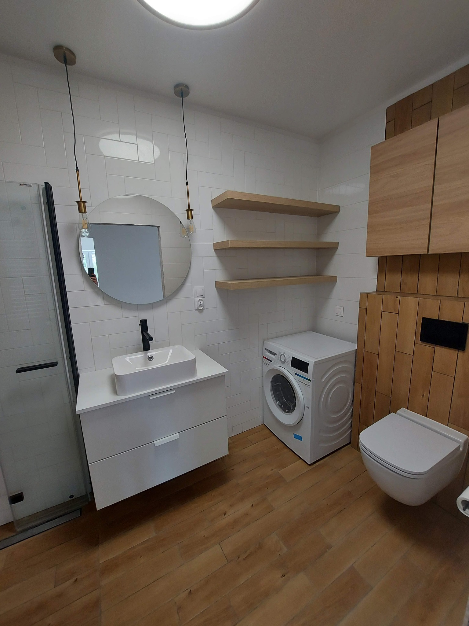 Nowoczesna łazienka z białymi płytkami, drewnianą podłogą i akcentami z drewna na ścianie, okrągłe lustro z wiszącymi lampami, biała umywalka nablatowa, wisząca toaleta i pralka.
