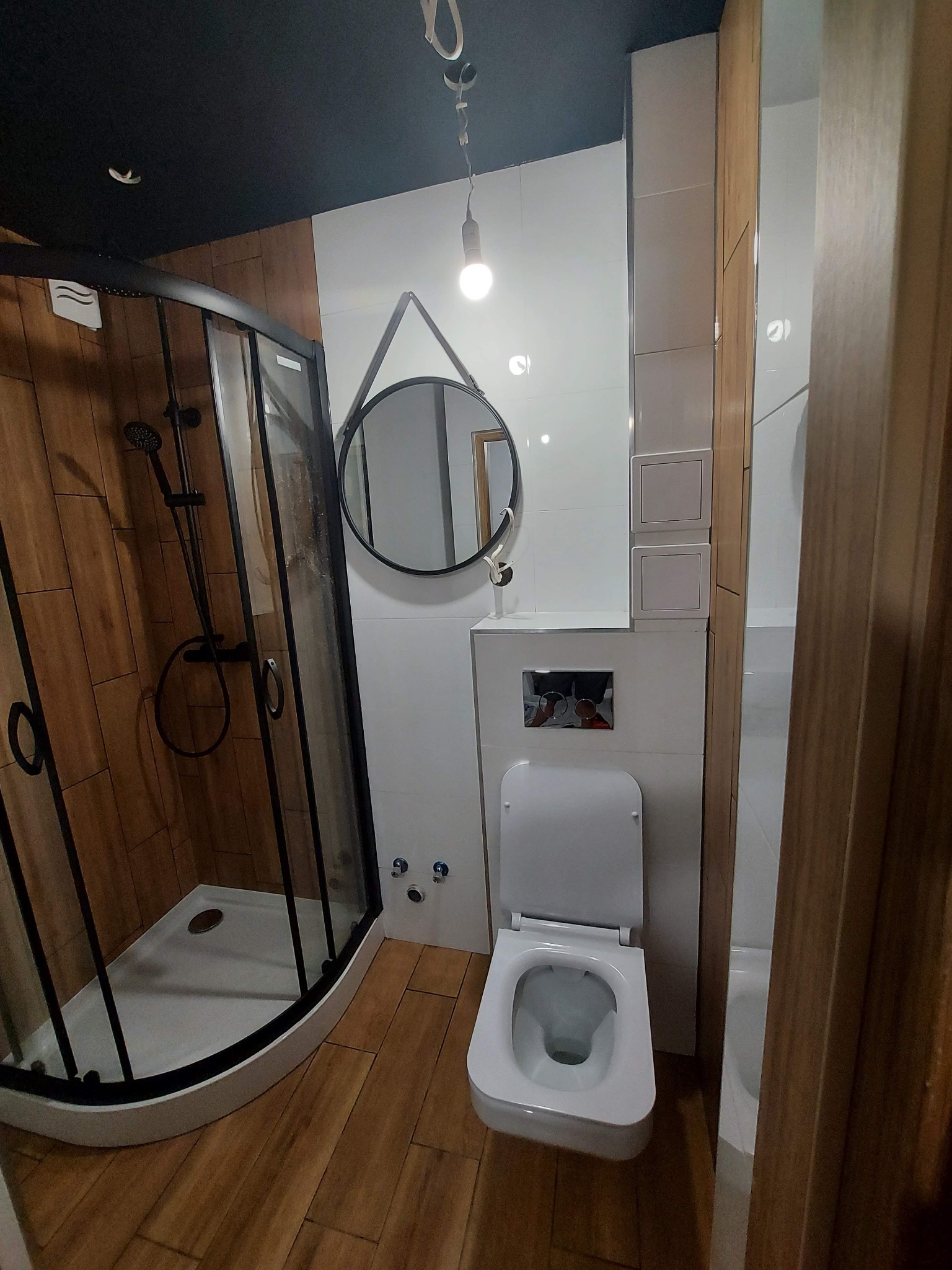 Nowoczesna łazienka z kabiną prysznicową w czarnej ramie, okrągłym lustrem wiszącym na ścianie i białymi płytkami, uzupełniona drewnopodobną podłogą i toaletą z podtynkowym systemem spłukiwania.