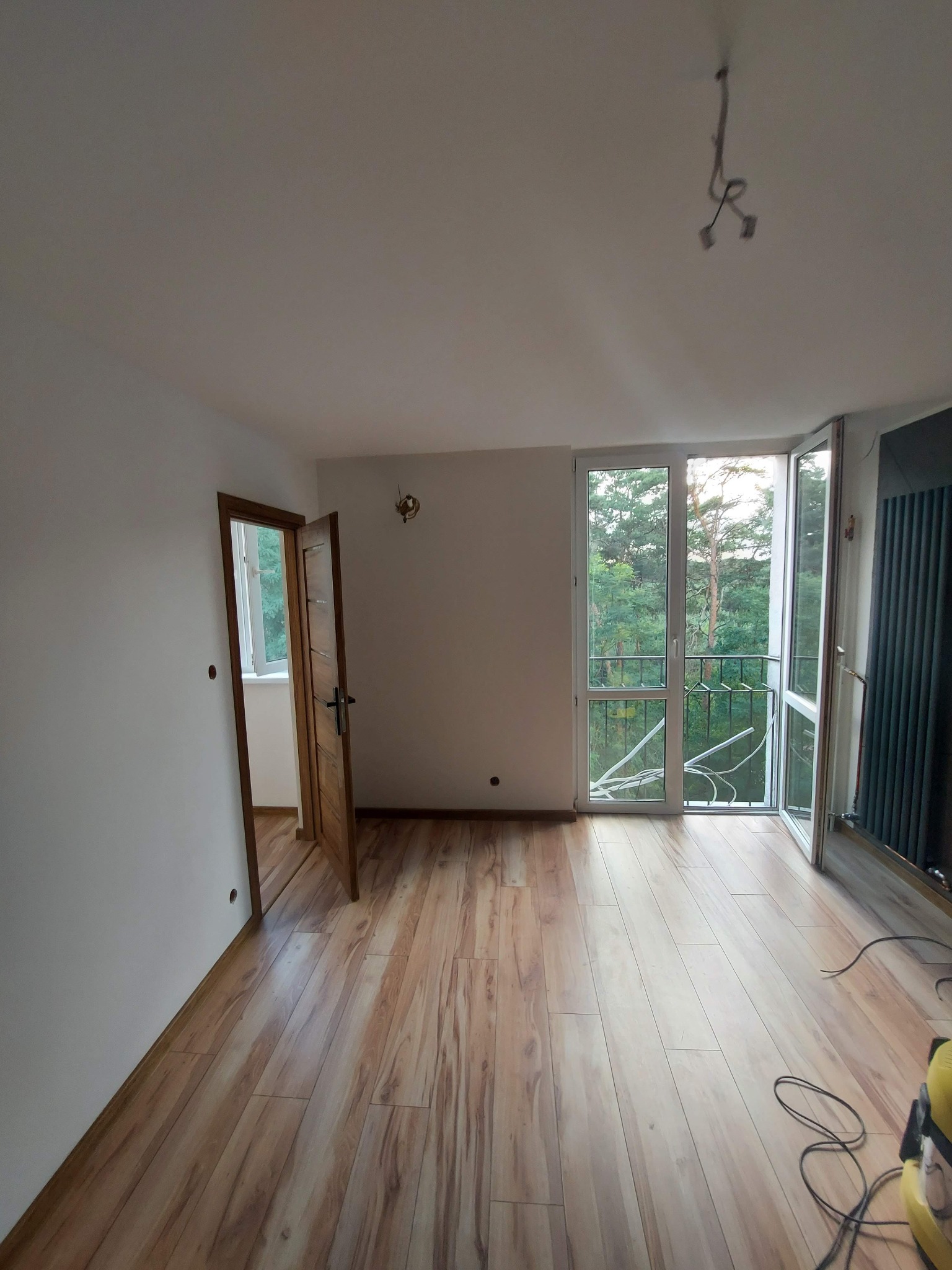 Jasne, przestronne wnętrze po remoncie z drewnianą podłogą, otwartymi drzwiami balkonowymi z widokiem na zieleń, i drewnianymi drzwiami wewnętrznymi, z widocznymi przewodami elektrycznymi...