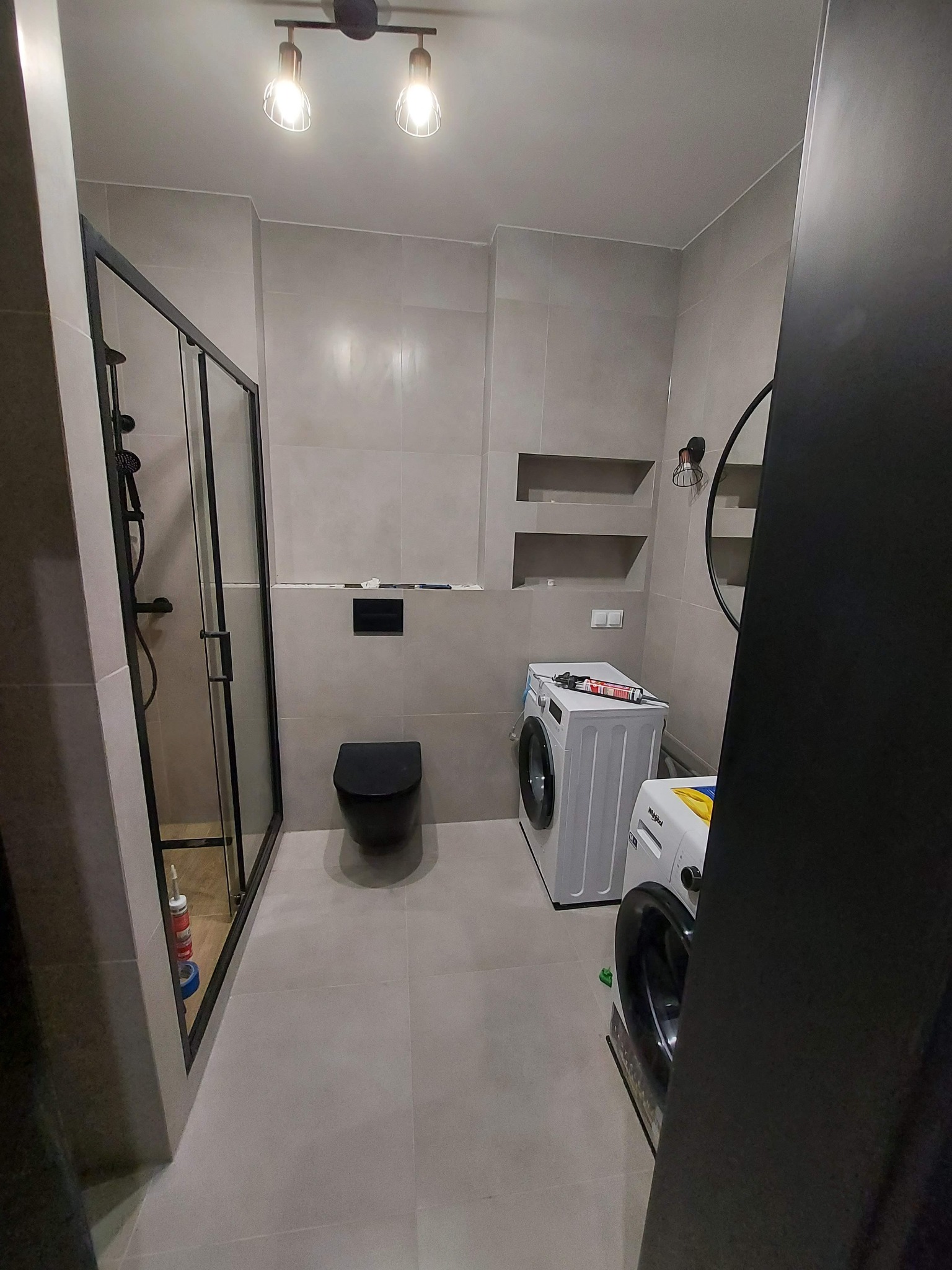 Wykończona łazienka w Bydgoszczy z kabiną prysznicową z czarnymi ramami, podwieszaną toaletą, dwiema pralkami i szarą glazurą na ścianach oraz sufitem z oświetleniem punktowym.