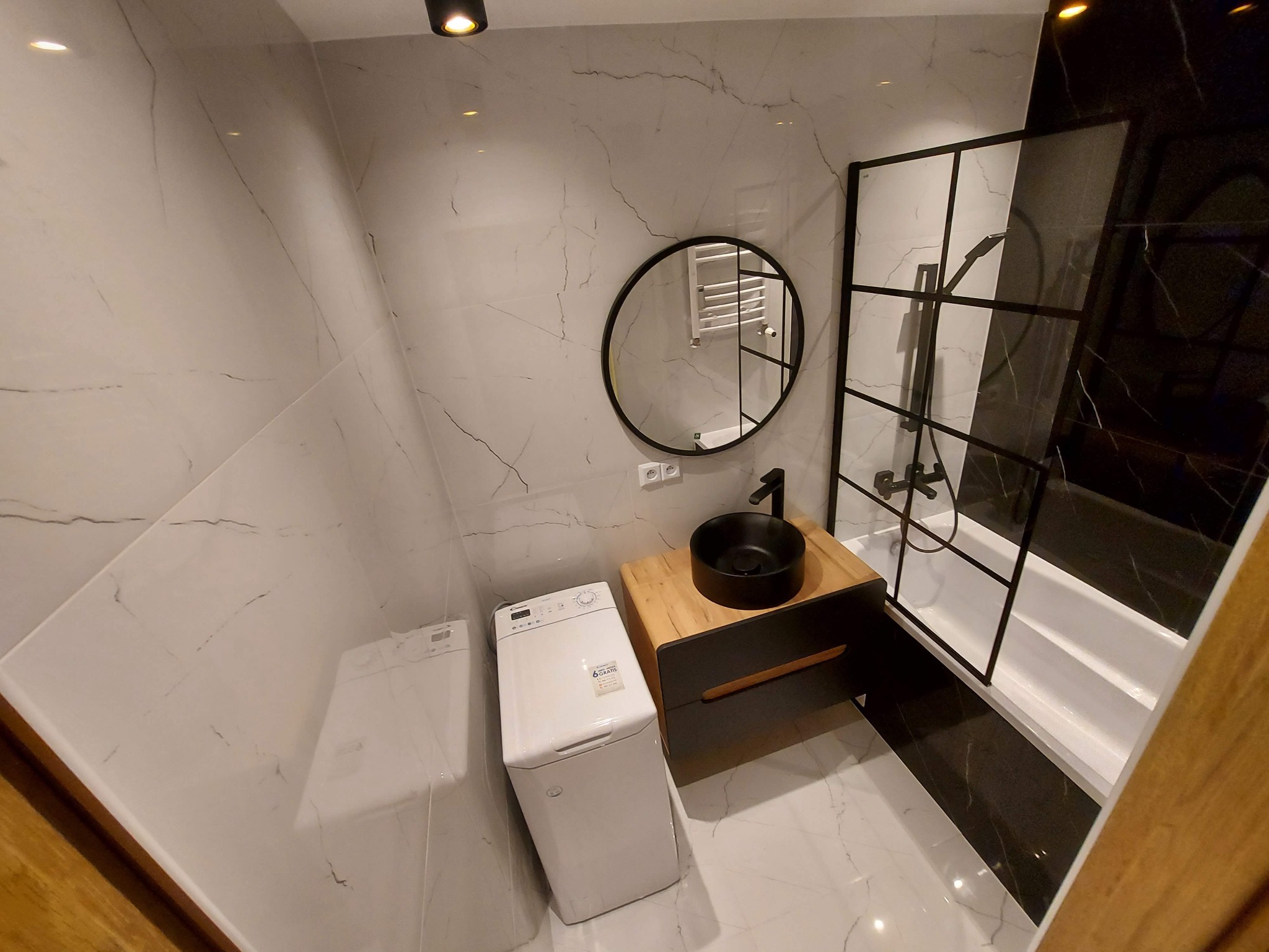 Nowoczesna łazienka z marmurowymi płytkami, okrągłym lustrem, czarną umywalką nablatową na drewnianej szafce, kabiną prysznicową typu walk-in z czarnymi profilami i pralką kompaktową w rogu.