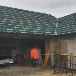 Budynek w trakcie budowy z częściowo wykonanym dachem pokrytym membraną i łatami, widoczny pracownik w pomarańczowej kurtce, materiały budowlane i narzędzia.