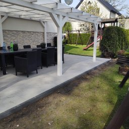 Companybruk - Nowoczesny taras z pergolą w ogrodzie. Szare płyty tarasowe, czarne meble ogrodowe, biała konstrukcja pergoli. W tle trawnik, huśtawka i dom.