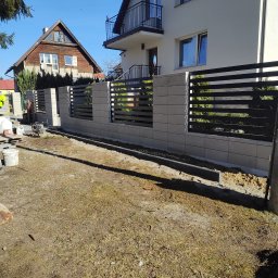 Companybruk - Nowoczesne ogrodzenie z szarej kostki i czarnych paneli poziomych, w trakcie montażu przed domem jednorodzinnym. Widoczne narzędzia i materiały budowlane.