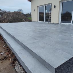 Companybruk - Ukończony taras z szarych płyt z dwoma stopniami prowadzącymi do domu z dużymi oknami. Widoczny fragment elewacji i sterta ziemi w tle.