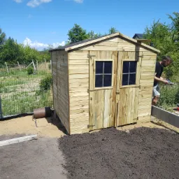 Nowy, drewniany domek narzędziowy z dwuskrzydłowymi drzwiami i dwoma oknami, zlokalizowany w ogrodzie na tle błękitnego nieba z chmurami.