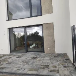 Nowoczesny taras z kostki brukowej w odcieniach szarości, przylegający do białego budynku z dużymi oknami. Widoczne elementy wykończeniowe i fragment ogrodzenia.