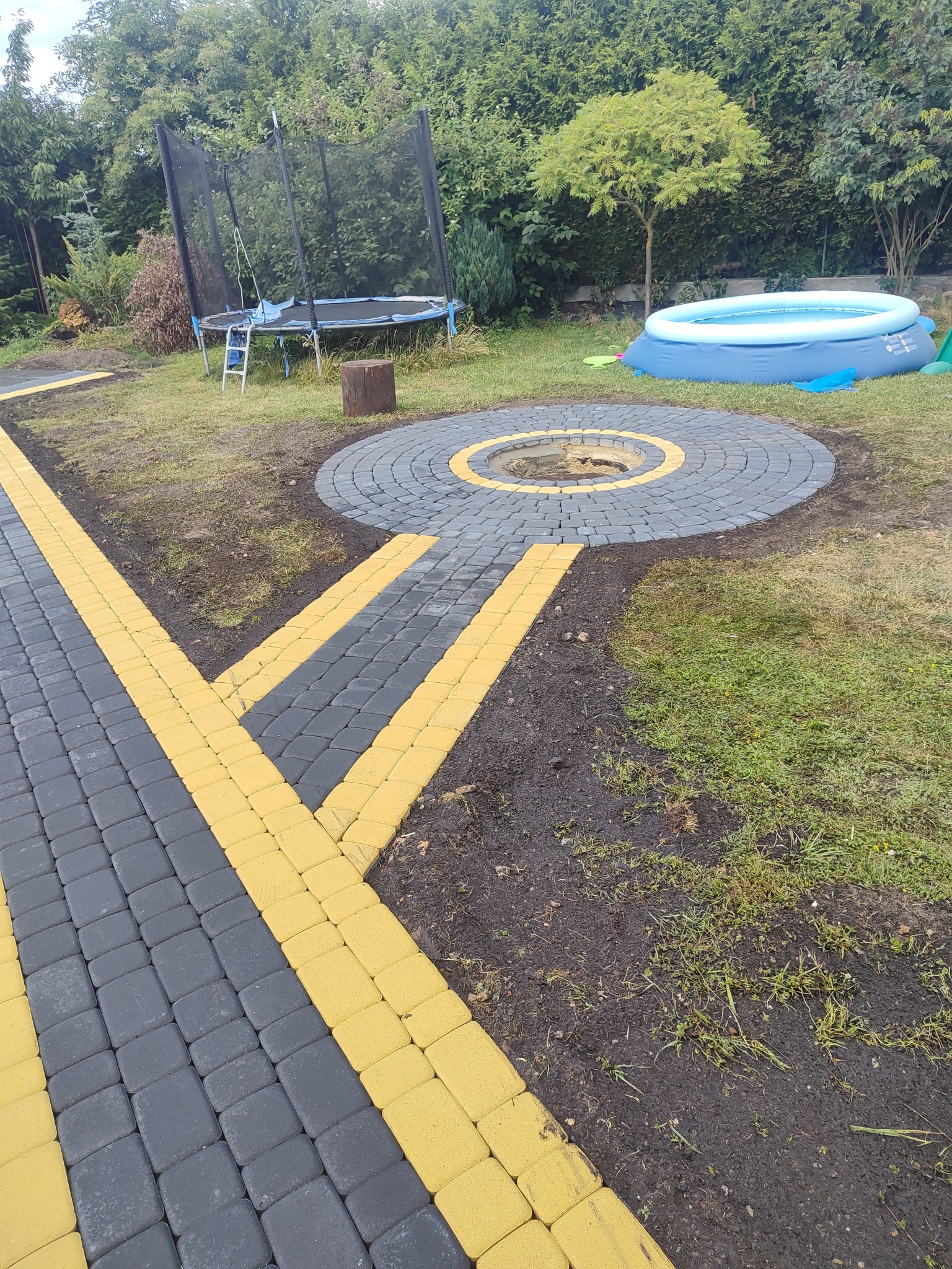 Ścieżka z kostki brukowej w kolorze szaro-żółtym prowadząca do okrągłego paleniska na trawniku, w tle trampolina i basen.