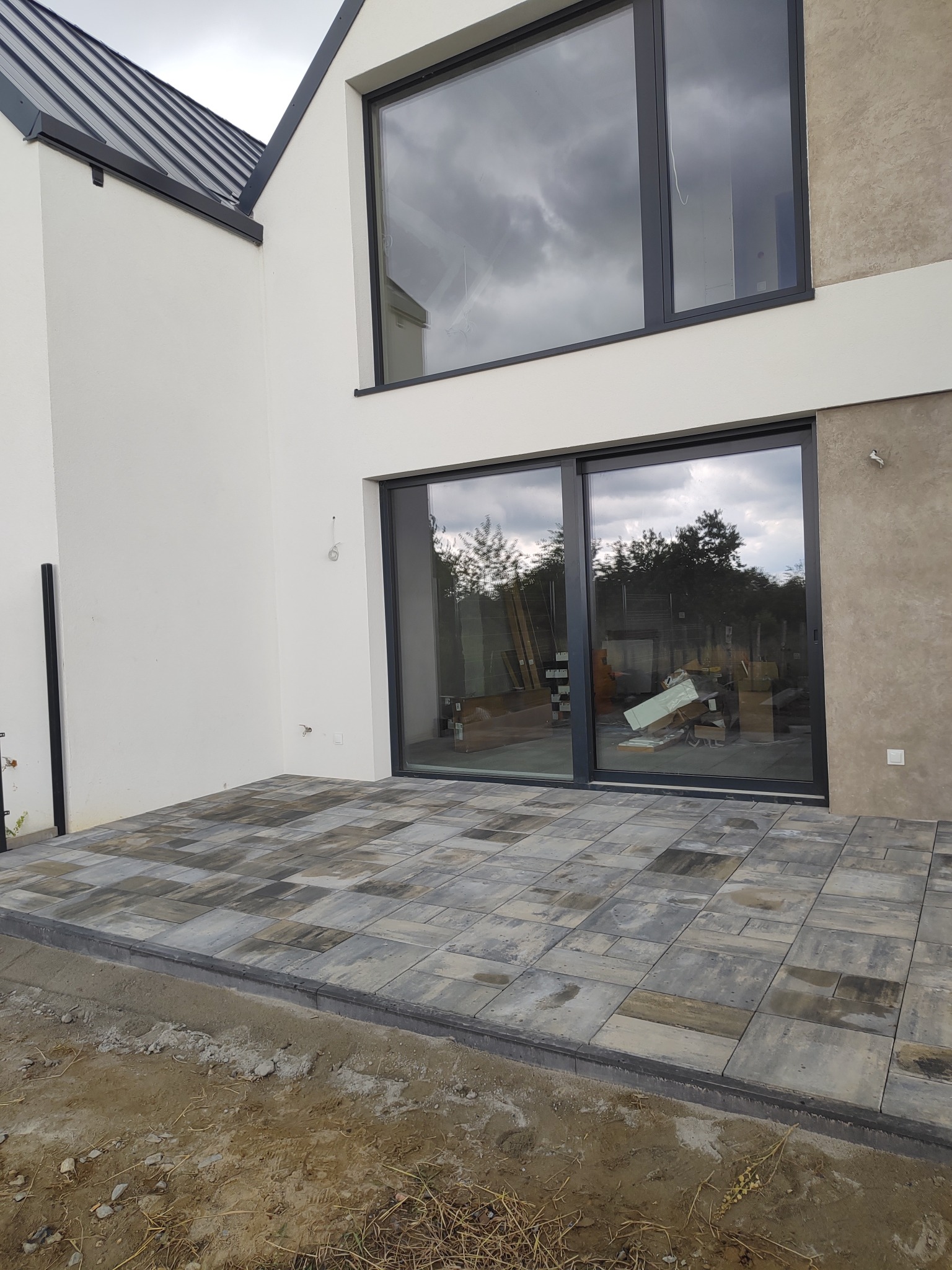 Nowoczesny taras z kostki brukowej w odcieniach szarości, przylegający do budynku z dużymi oknami przesuwnymi. Widoczne niedokończone obrzeża tarasu.