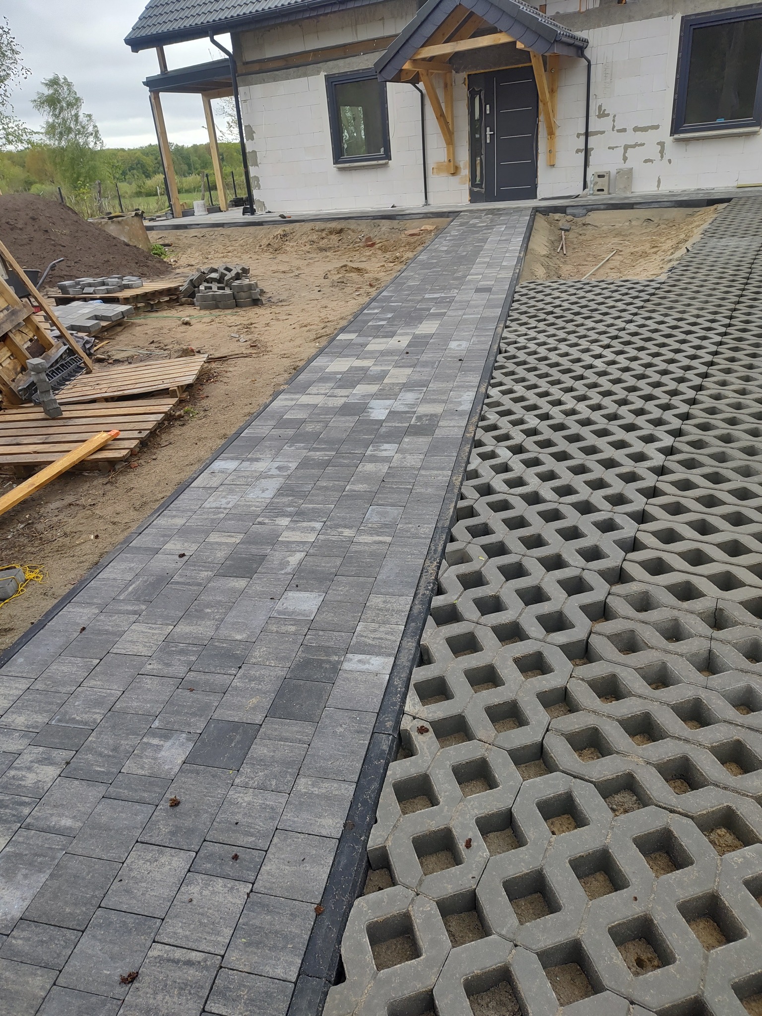 Ułożona ścieżka z szarej kostki brukowej prowadząca do domu, obok ażurowa kostka trawnikowa. Widoczne materiały budowlane i fragment elewacji budynku.