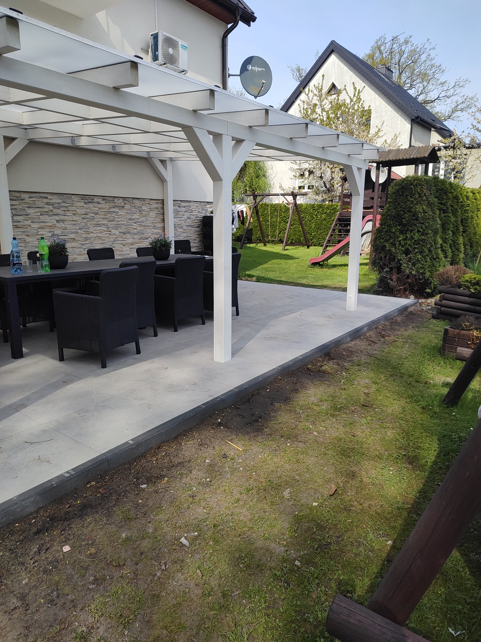 Nowoczesny taras z pergolą w ogrodzie. Szare płyty tarasowe, czarne meble ogrodowe, biała konstrukcja pergoli. W tle trawnik, huśtawka i dom.