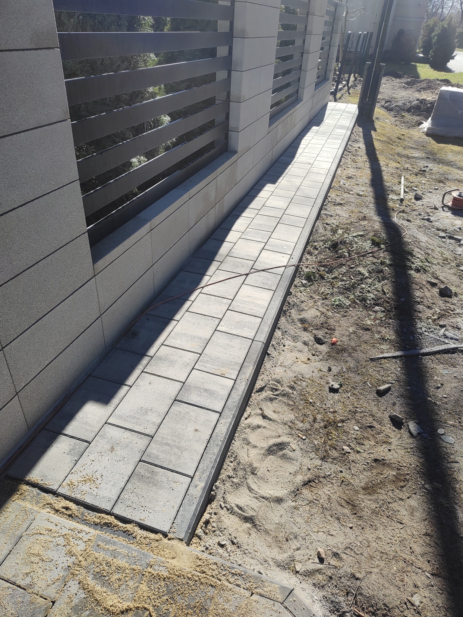 Ścieżka z szarych kostek brukowych ułożona wzdłuż nowoczesnego, szarego ogrodzenia z poziomymi, czarnymi przęsłami. Widoczne ślady piasku i ziemi.