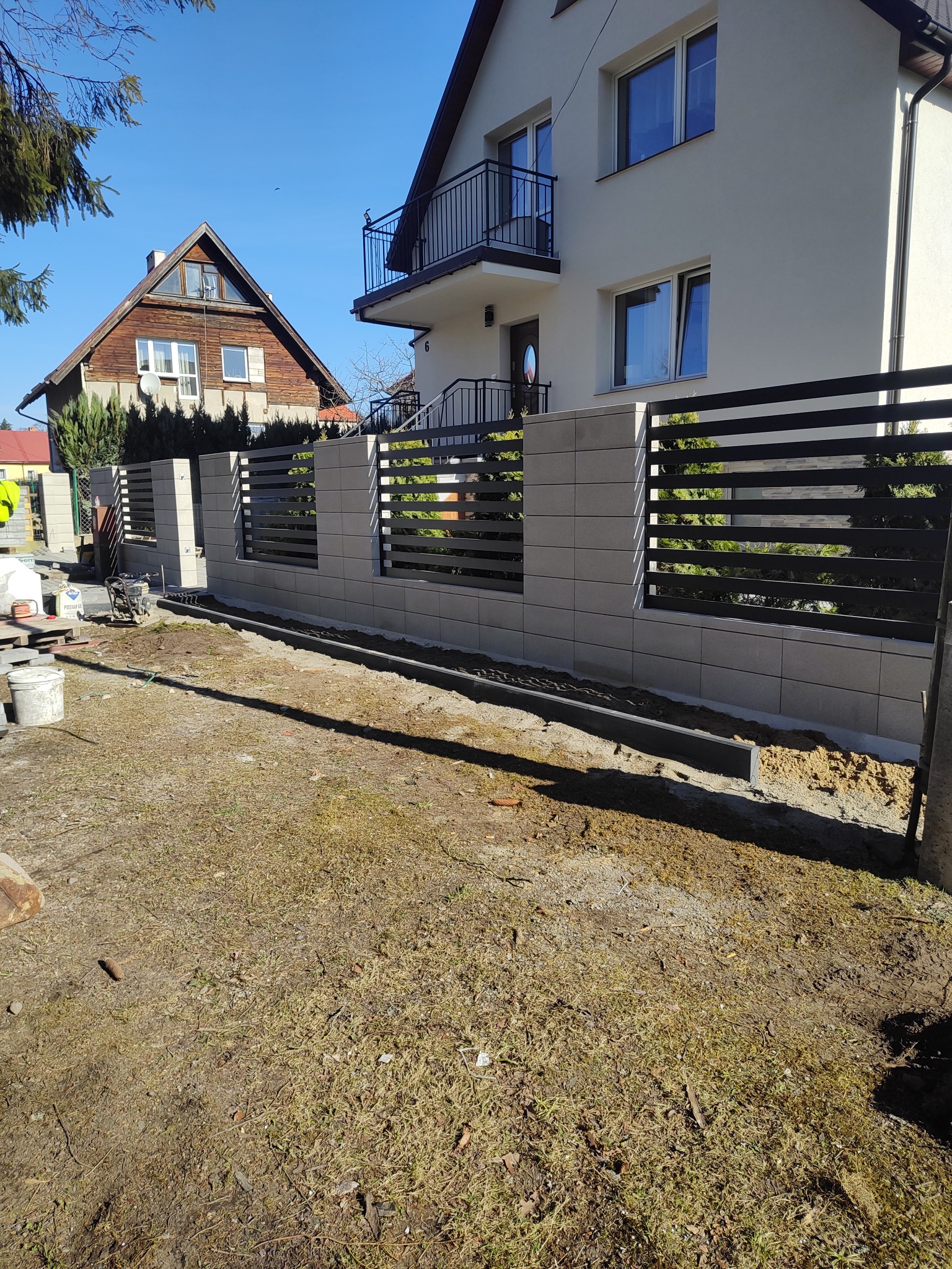 Nowoczesne ogrodzenie z szarej kostki i czarnych paneli poziomych, w trakcie montażu przed domem jednorodzinnym. Widoczne narzędzia i materiały budowlane.