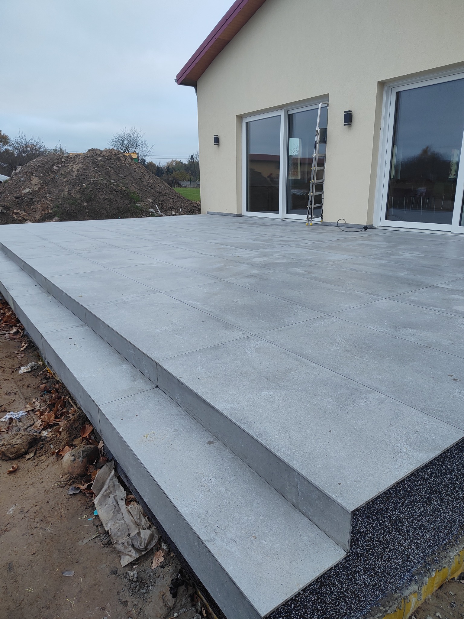 Ukończony taras z szarych płyt z dwoma stopniami prowadzącymi do domu z dużymi oknami. Widoczny fragment elewacji i sterta ziemi w tle.