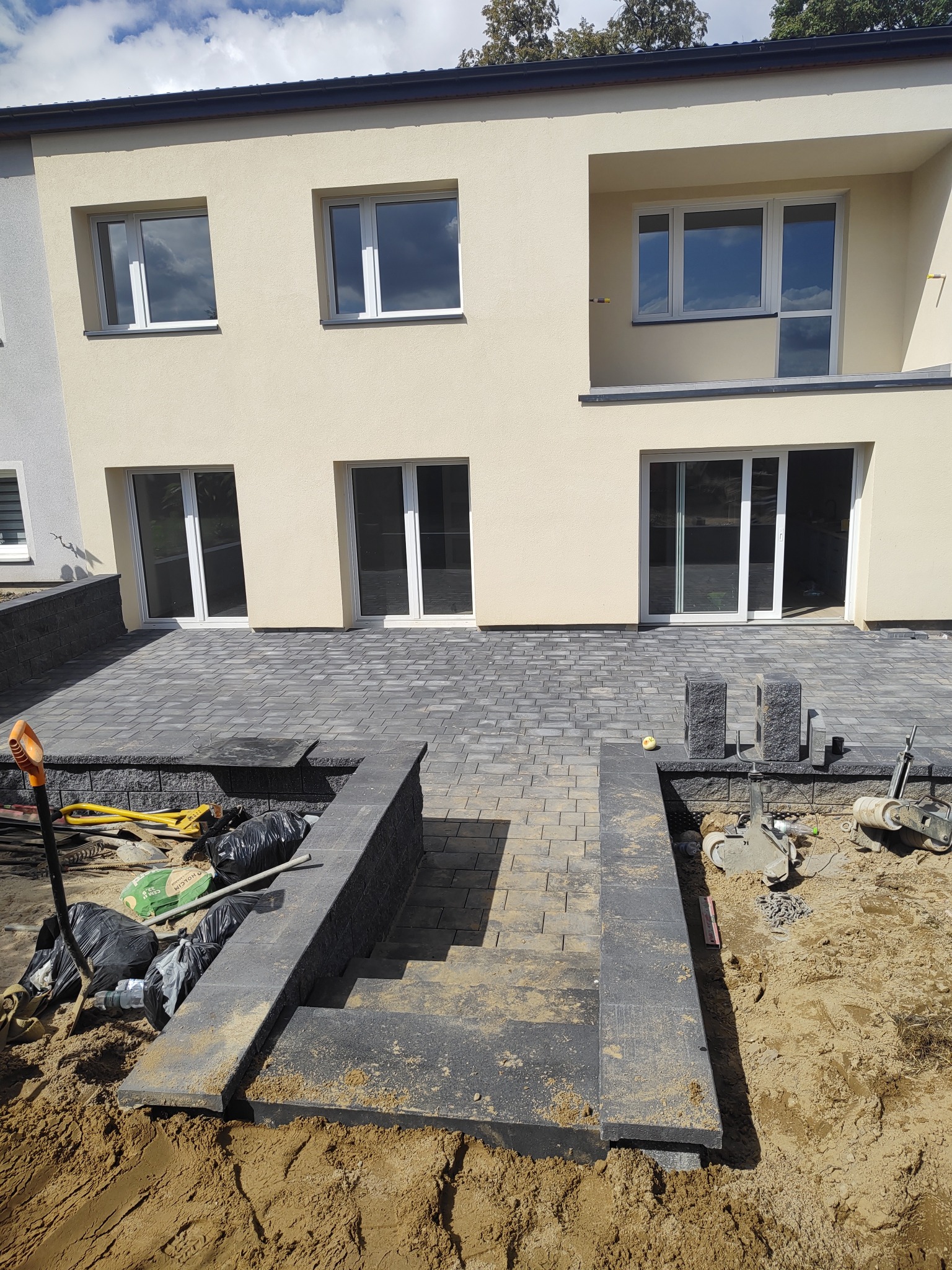 Schody z kostki brukowej prowadzące do tarasu przy domu. Murki oporowe, częściowo wykonane. Widoczne narzędzia i materiały budowlane.