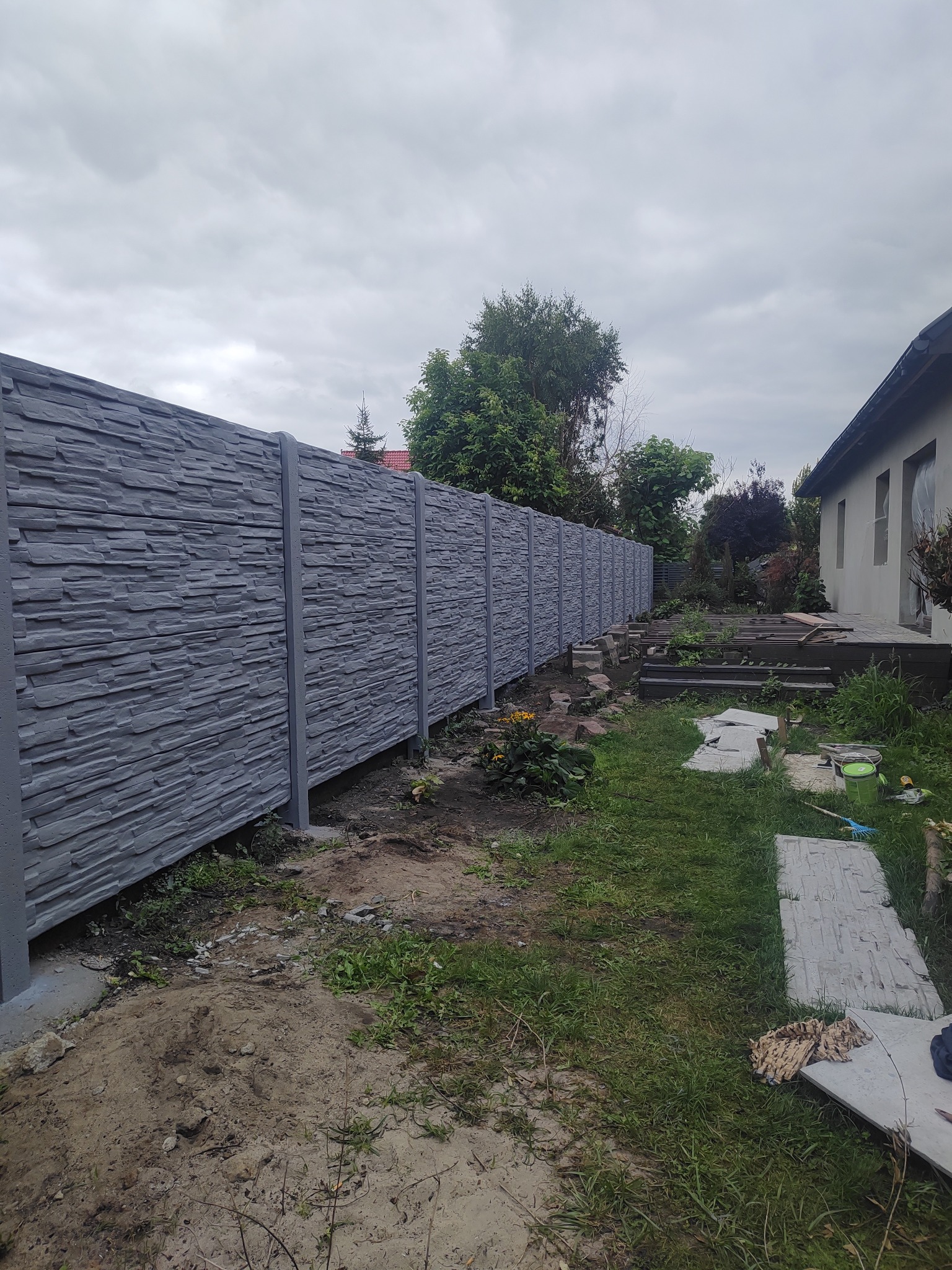 Świeżo pomalowany, podwójnie opłytowany płot betonowy w kolorze popiel, z fakturą cegły, widok z boku na tle zieleni i fragmentu budynku.