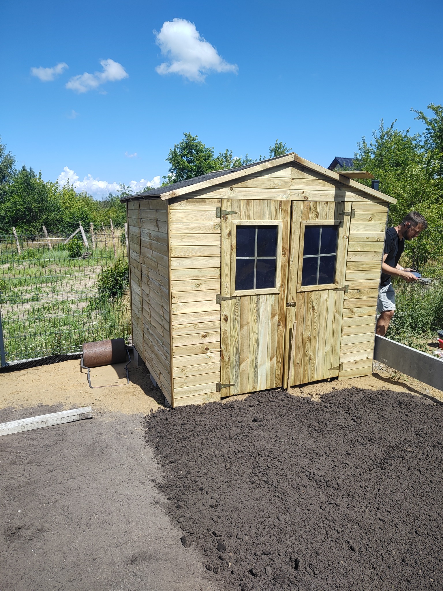 Nowy, drewniany domek narzędziowy z dwuskrzydłowymi drzwiami i dwoma oknami, zlokalizowany w ogrodzie na tle błękitnego nieba z chmurami.