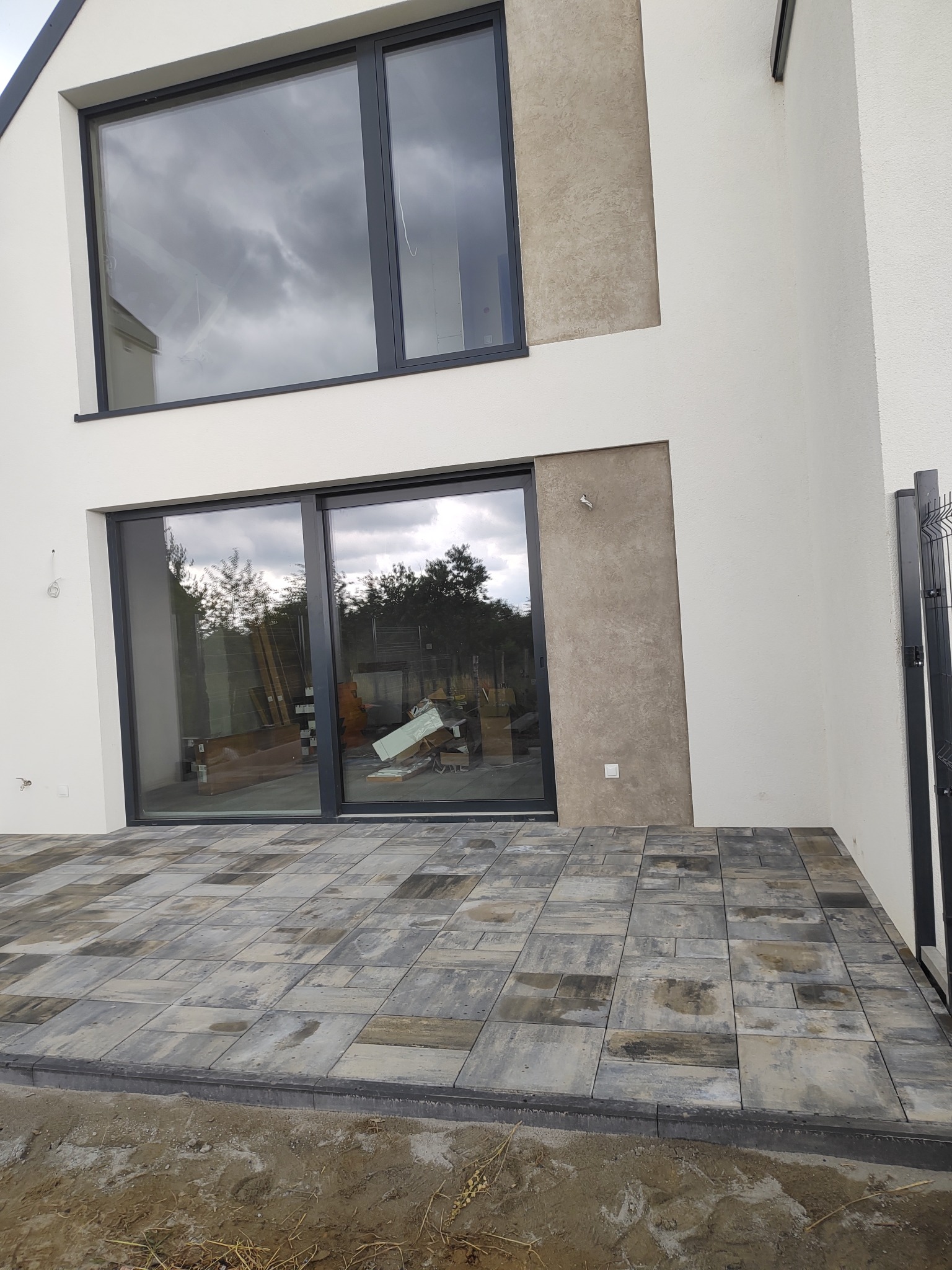 Nowoczesny taras z kostki brukowej w odcieniach szarości, przylegający do białego budynku z dużymi oknami. Widoczne elementy wykończeniowe i fragment ogrodzenia.