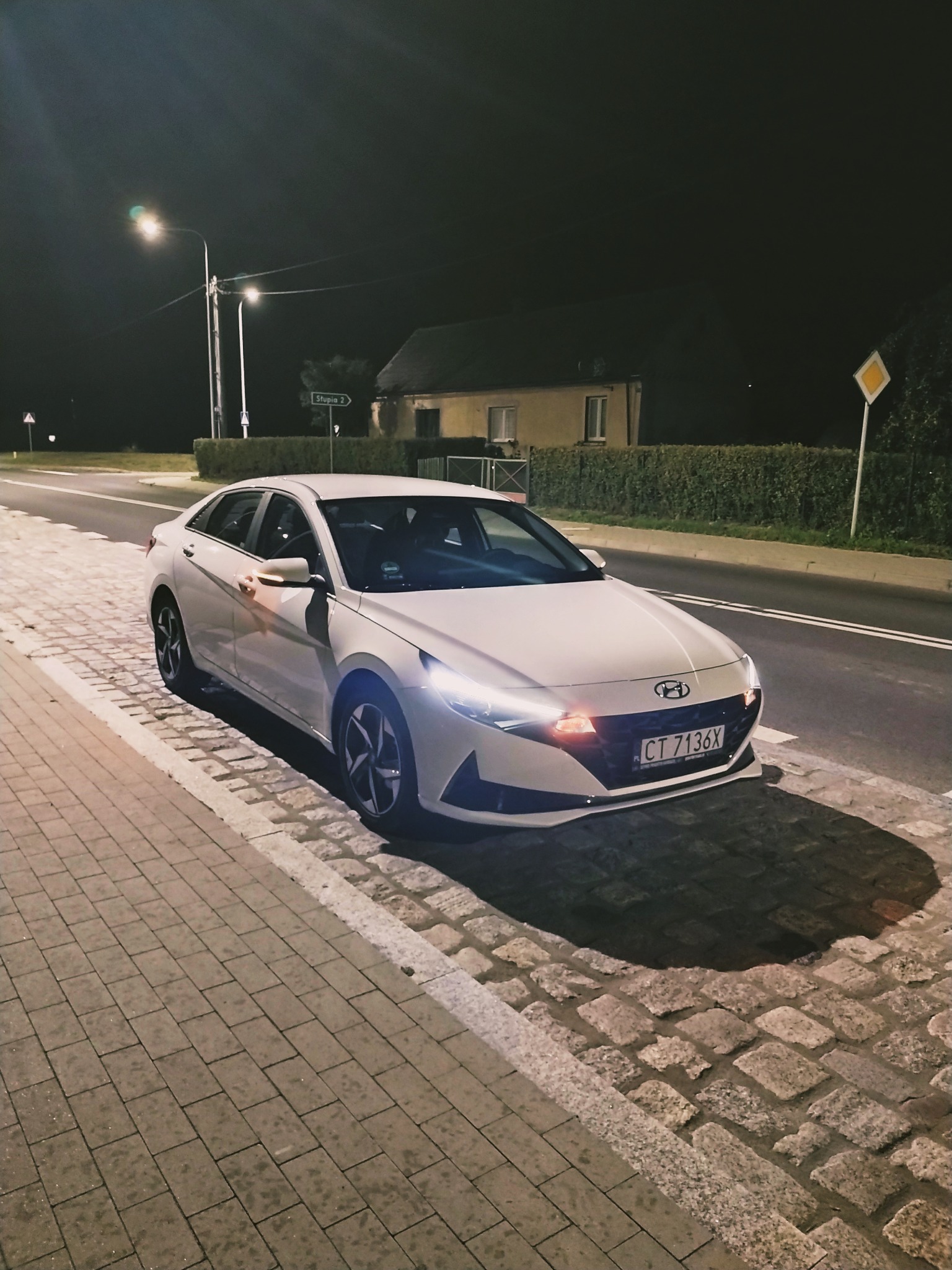 Biały samochód osobowy Hyundai zaparkowany nocą przy oświetlonej ulicy z kostki brukowej, widoczny znak drogowy D-6.