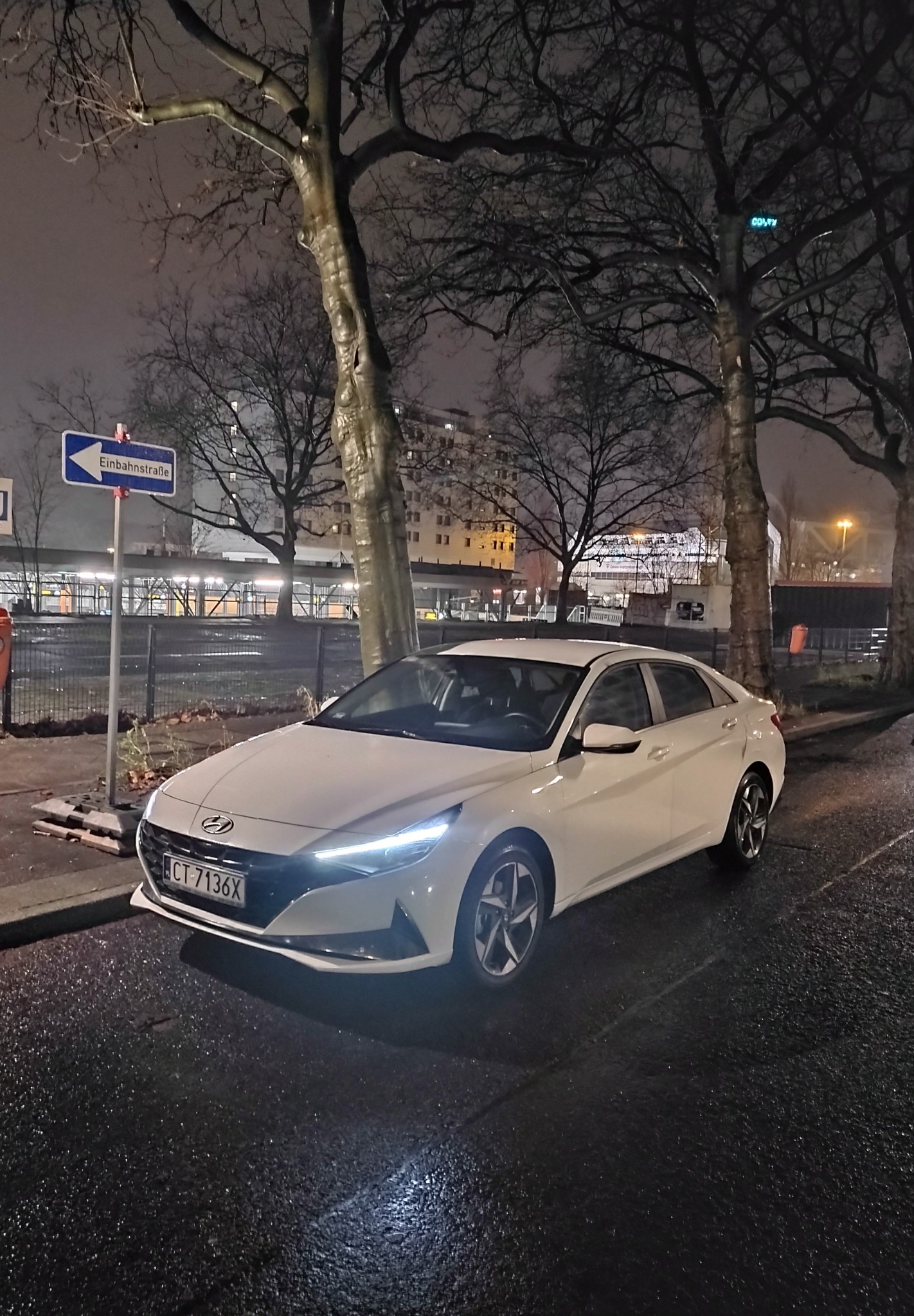 Biały samochód Hyundai Elantra zaparkowany na mokrej ulicy w nocy, widoczny znak drogowy 'Einbahnstraße' i budynki w tle.
