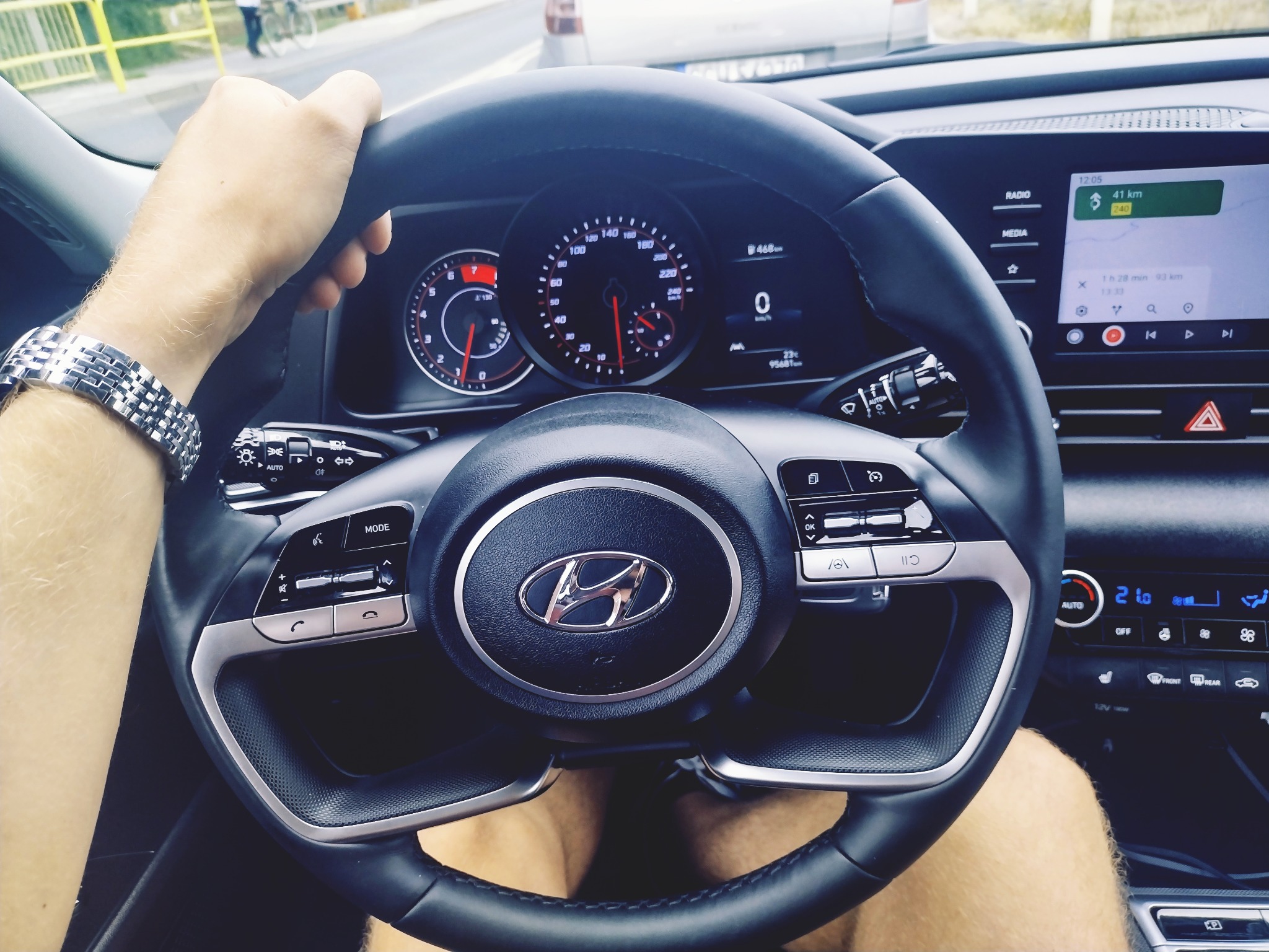 Wnętrze nowoczesnego samochodu z widocznym kierowcą, zegary z czerwonymi wskazówkami, nawigacja ustawiona na 41 km, zegarek na ręce kierowcy.