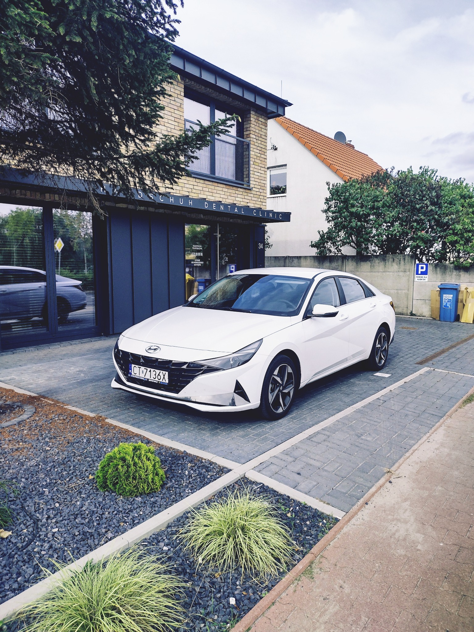 Biały sedan Hyundai zaparkowany przed nowoczesnym budynkiem z napisem 'DSCHUH DENTAL CLINIC' i numerem 34, obok znaku parkingowego i kosza na papier.