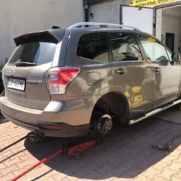 Szary SUV Subaru Forester, podniesiony na lewarku, zdemontowane tylne koło, widoczny warsztat w tle.