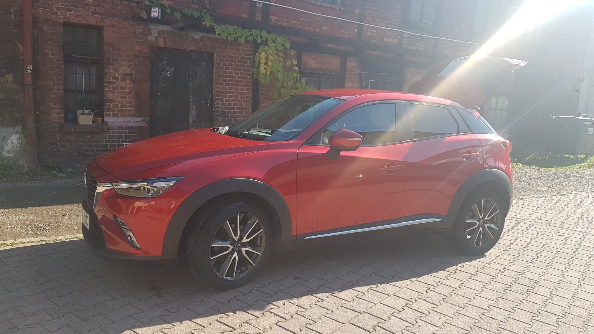 Czerwona Mazda CX-3 zaparkowana na brukowanej powierzchni przed ceglanym budynkiem z otwartą klapą bagażnika, oświetlona silnym słońcem.