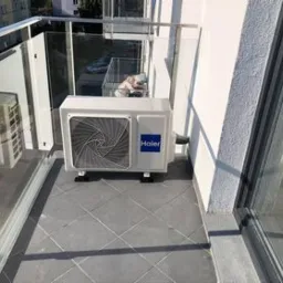 Biała jednostka zewnętrzna klimatyzacji Haier zamontowana na szarym balkonie z przeszkloną balustradą, widok z góry.