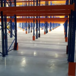 Długi rząd niebieskich metalowych słupk&oacute;w z pomarańczowymi podstawami i poprzeczkami, tworzących system regał&oacute;w w pustym magazynie z błyszczącą podłogą.