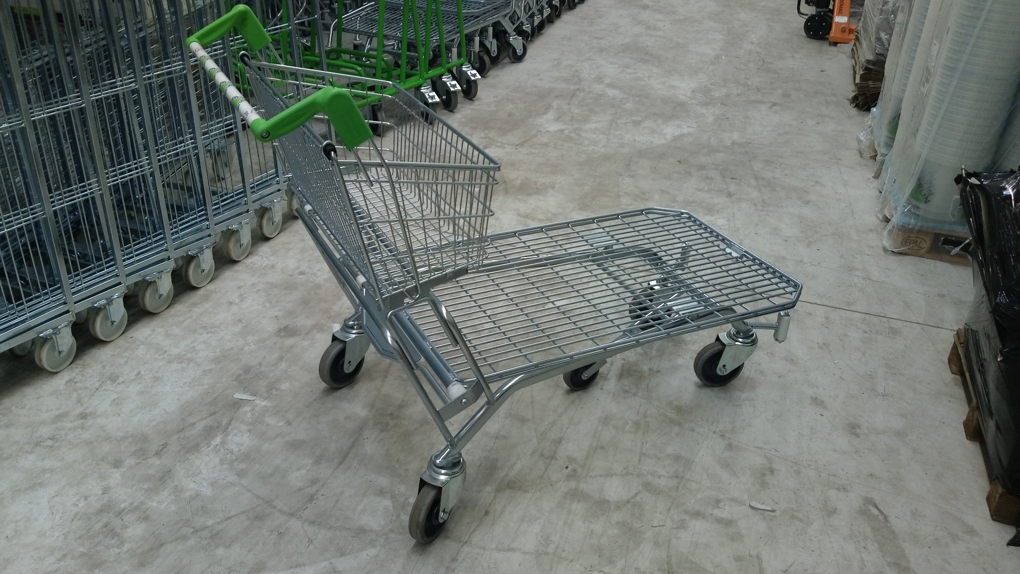 Wózek magazynowy z zielonymi uchwytami i metalową konstrukcją kosza oraz platformy, stojący na betonowej podłodze magazynu, w tle inne wózki i zapakowane towary.